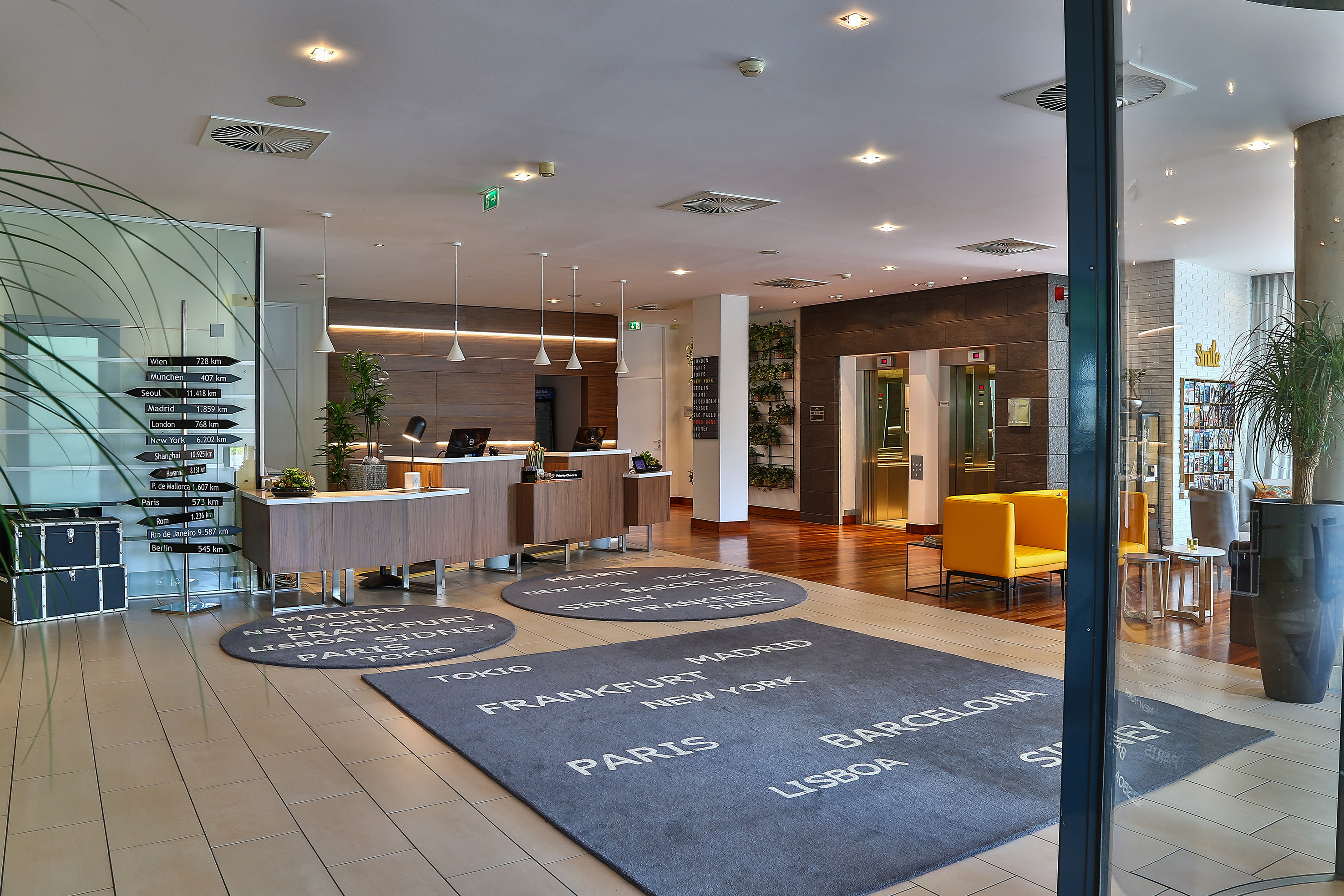Modern hotel lobby with reception, travel destination sign, and world city rugs.