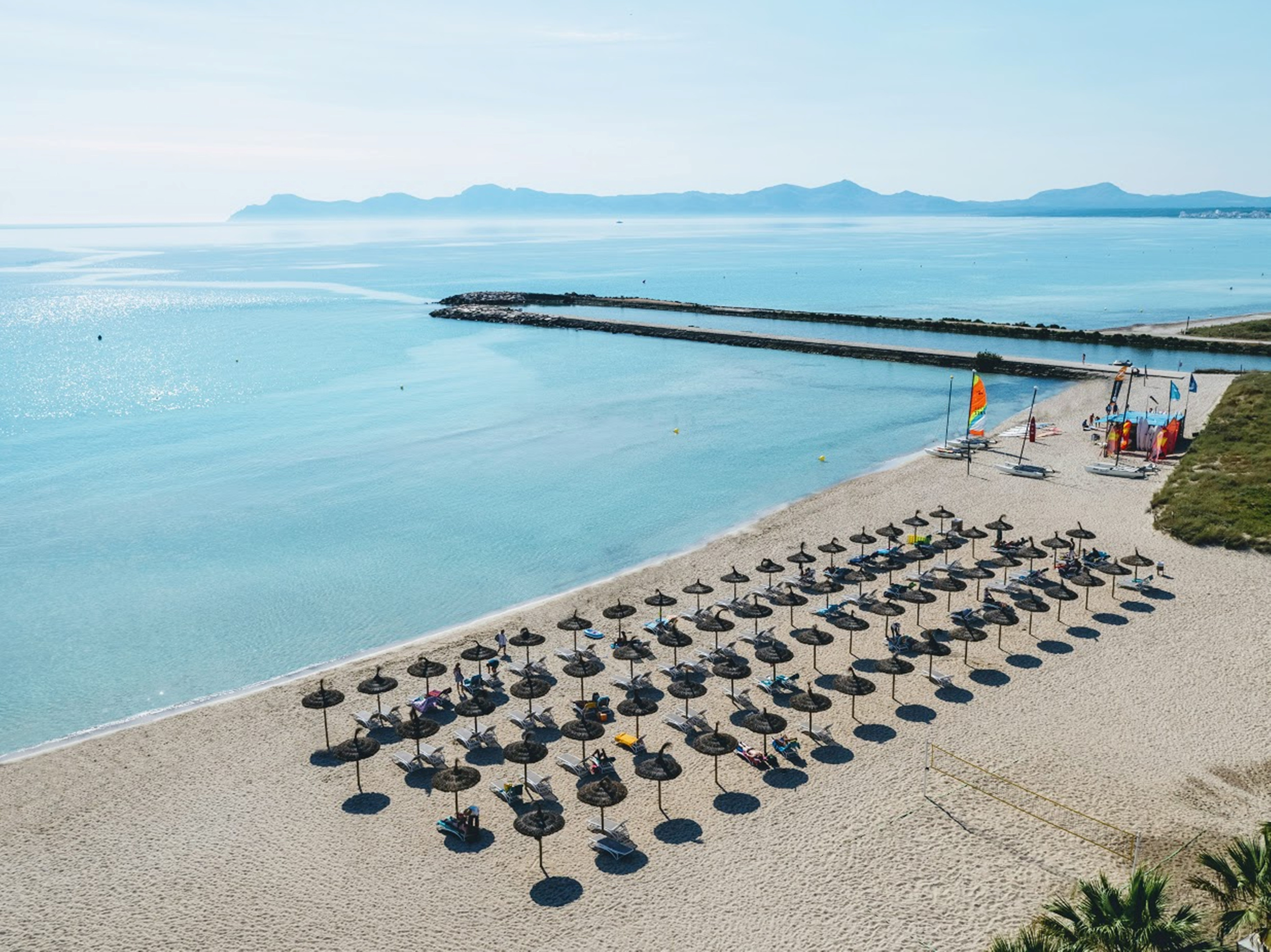 Playa arena, mar turquesa, sombrillas, muelle, barcos. Fondo montañoso.