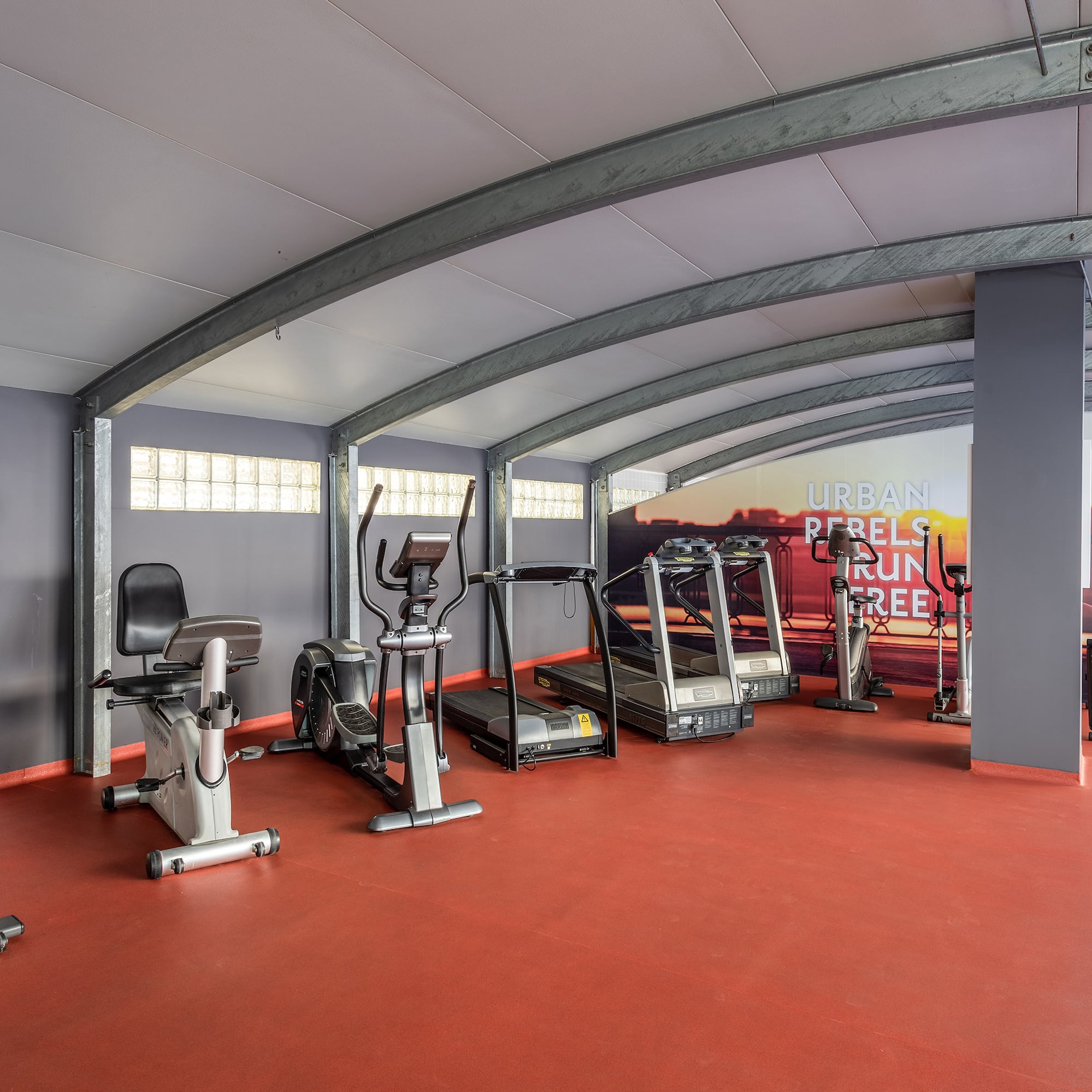 Spacious gym with treadmills, ellipticals, and stationary bikes on red flooring.