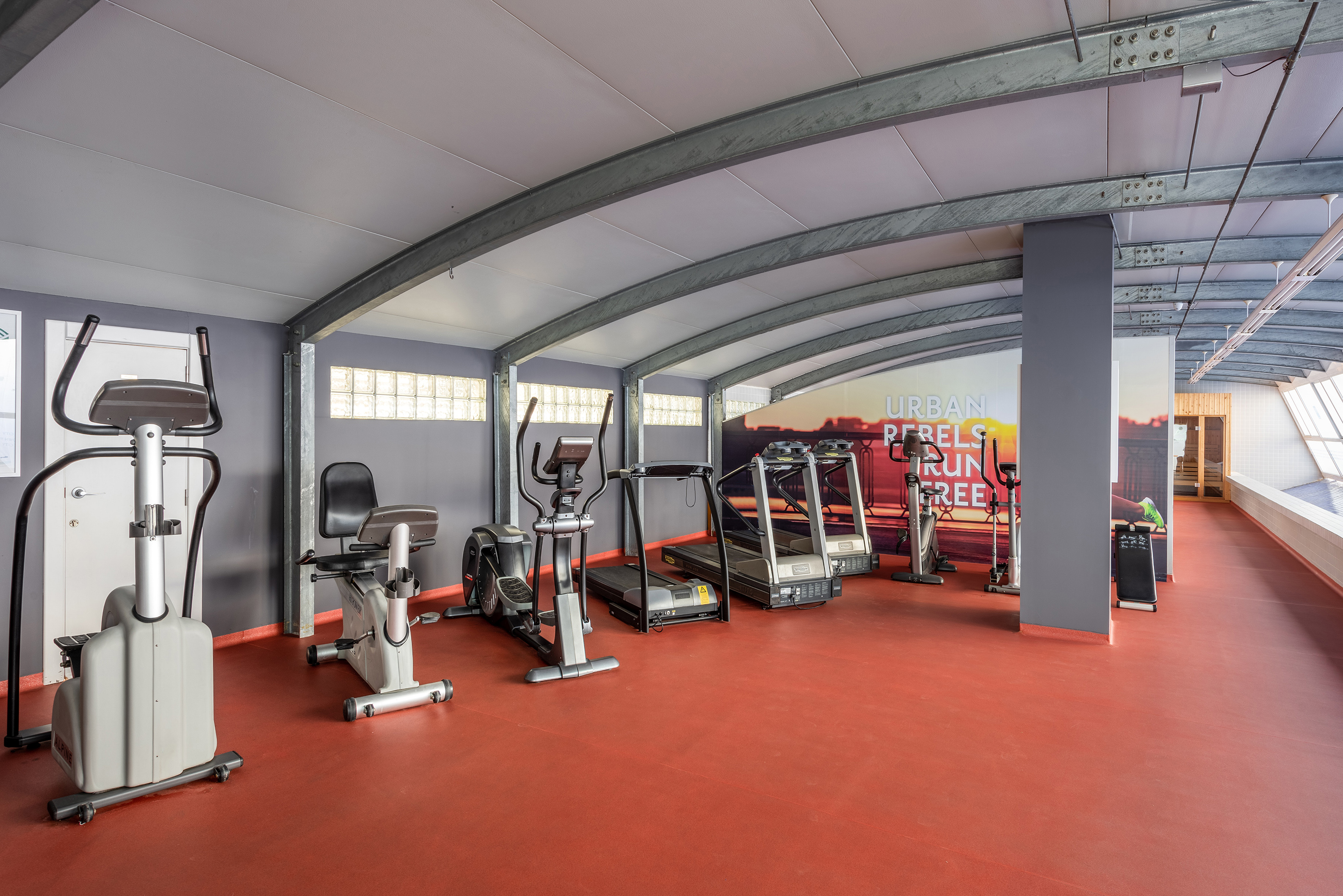 Spacious gym with treadmills, ellipticals, and stationary bikes on red flooring.