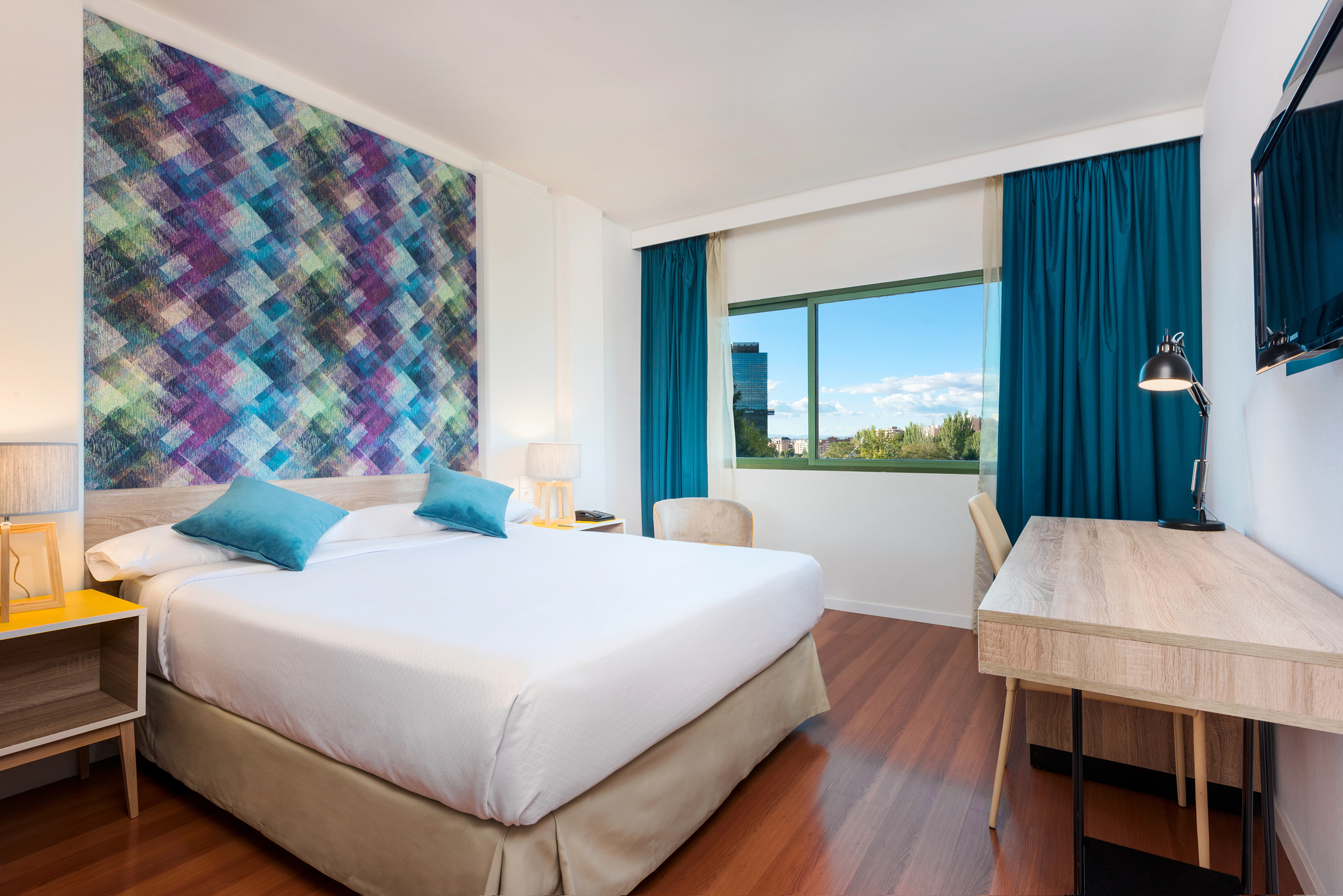 Modern hotel room with colorful geometric wall, large bed, and city view.