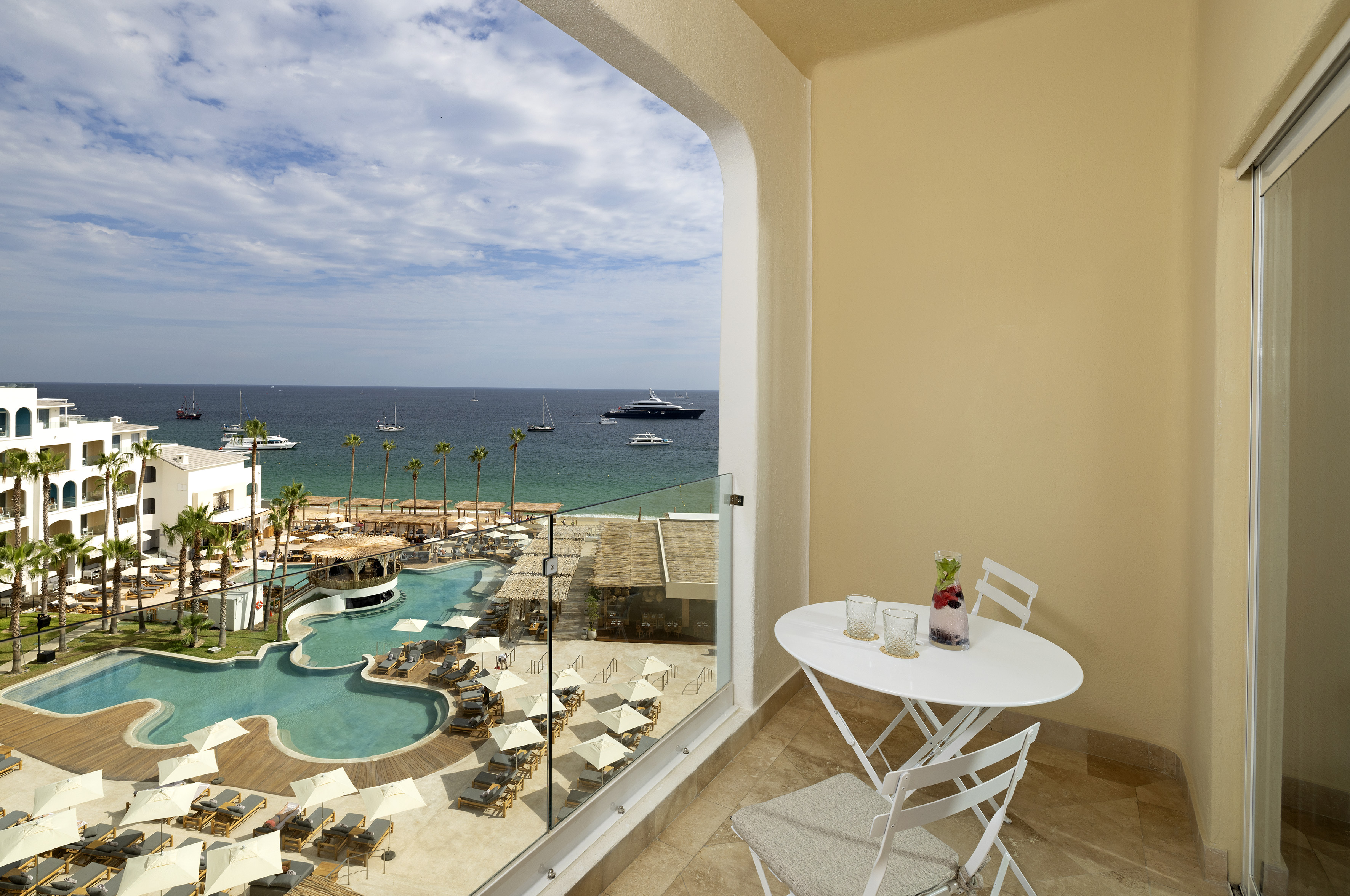 Luxury hotel balcony with ocean, resort pool, palm trees, and yachts.