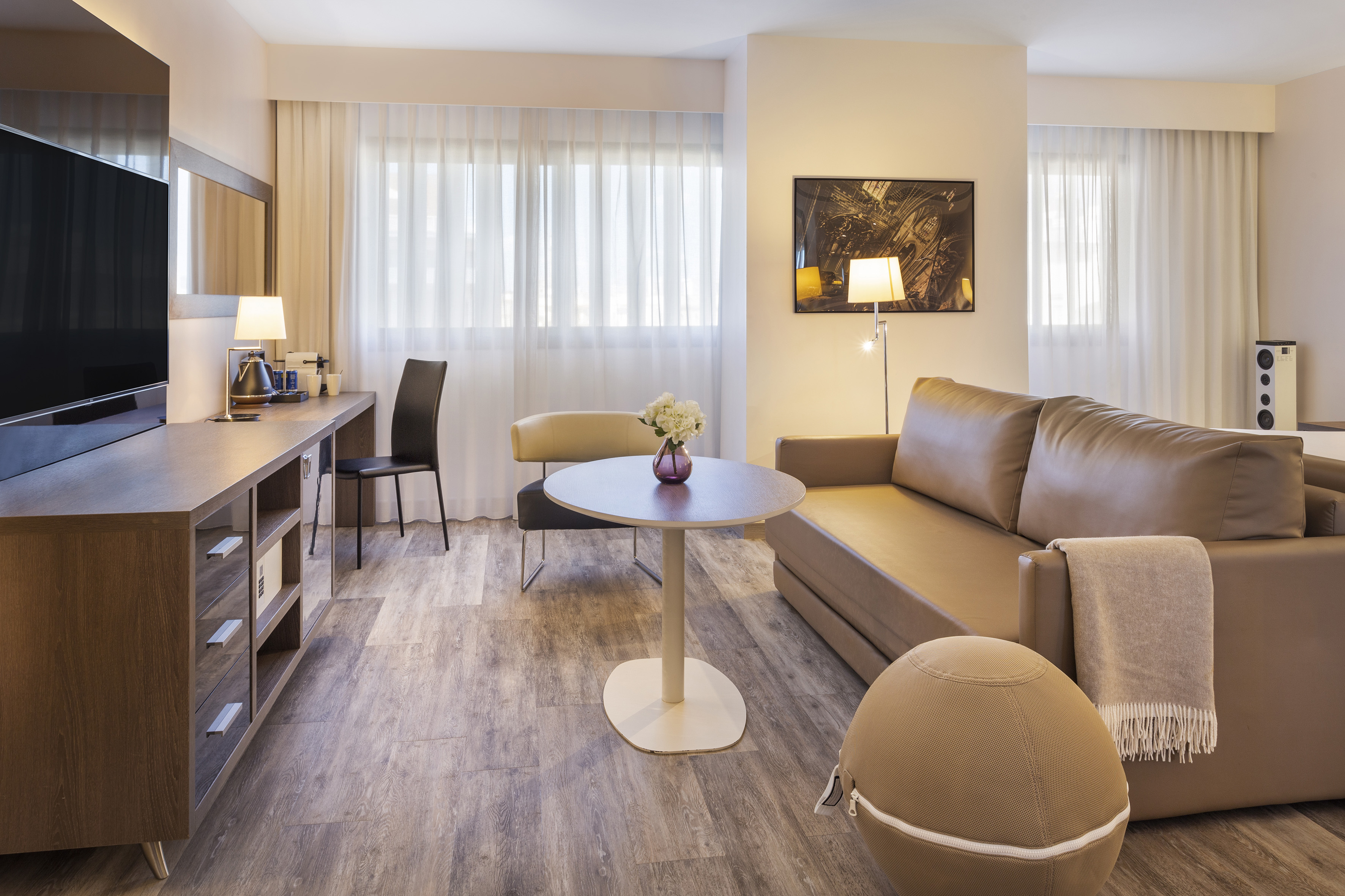 Modern hotel suite living area with tan sofa, wooden desk, TV, and coffee table.