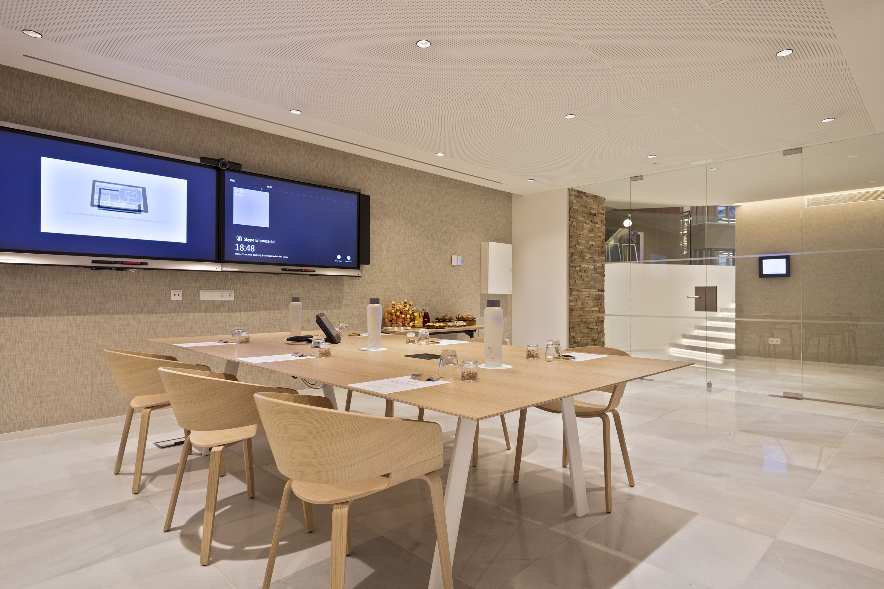 Modern conference room with dual screens for video conferencing, wooden table, and chairs.