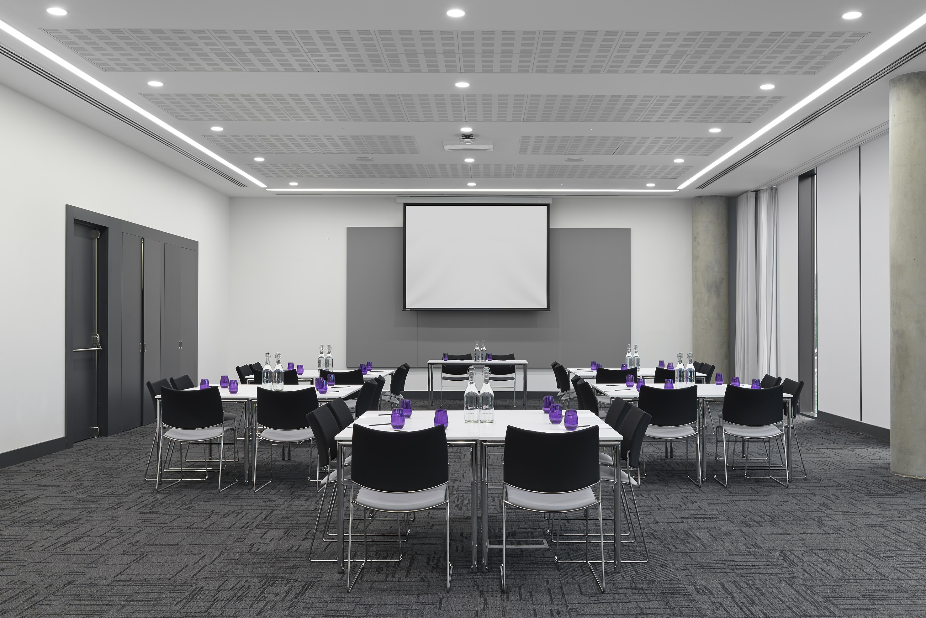 Empty contemporary meeting room with projector, tables, chairs, and patterned carpet.
