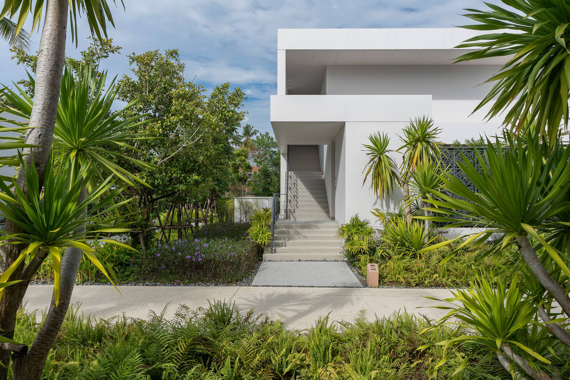 Modern white building with concrete stairs, lush tropical garden, and outdoor path.
