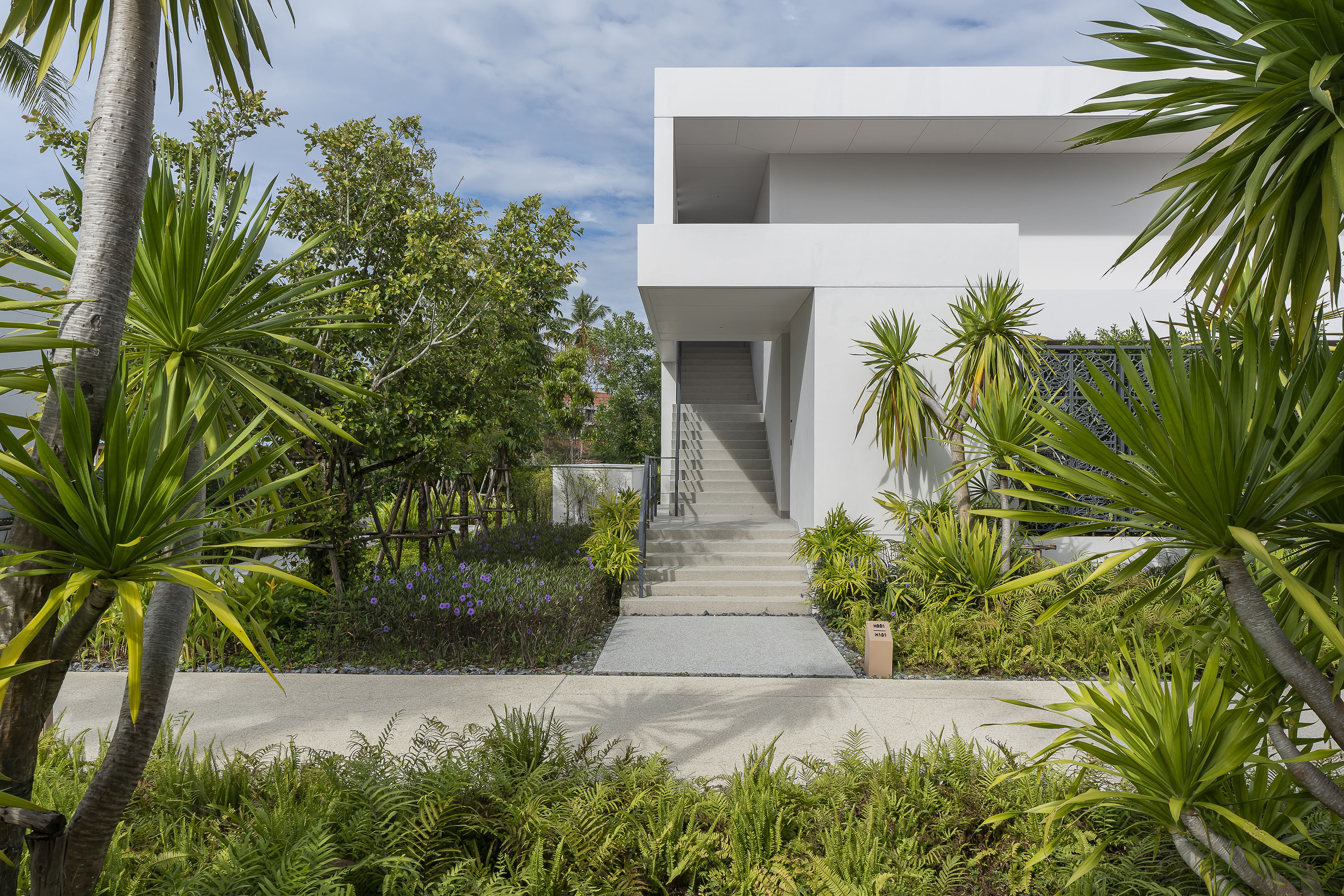 Modern white building with concrete stairs, lush tropical garden, and outdoor path.