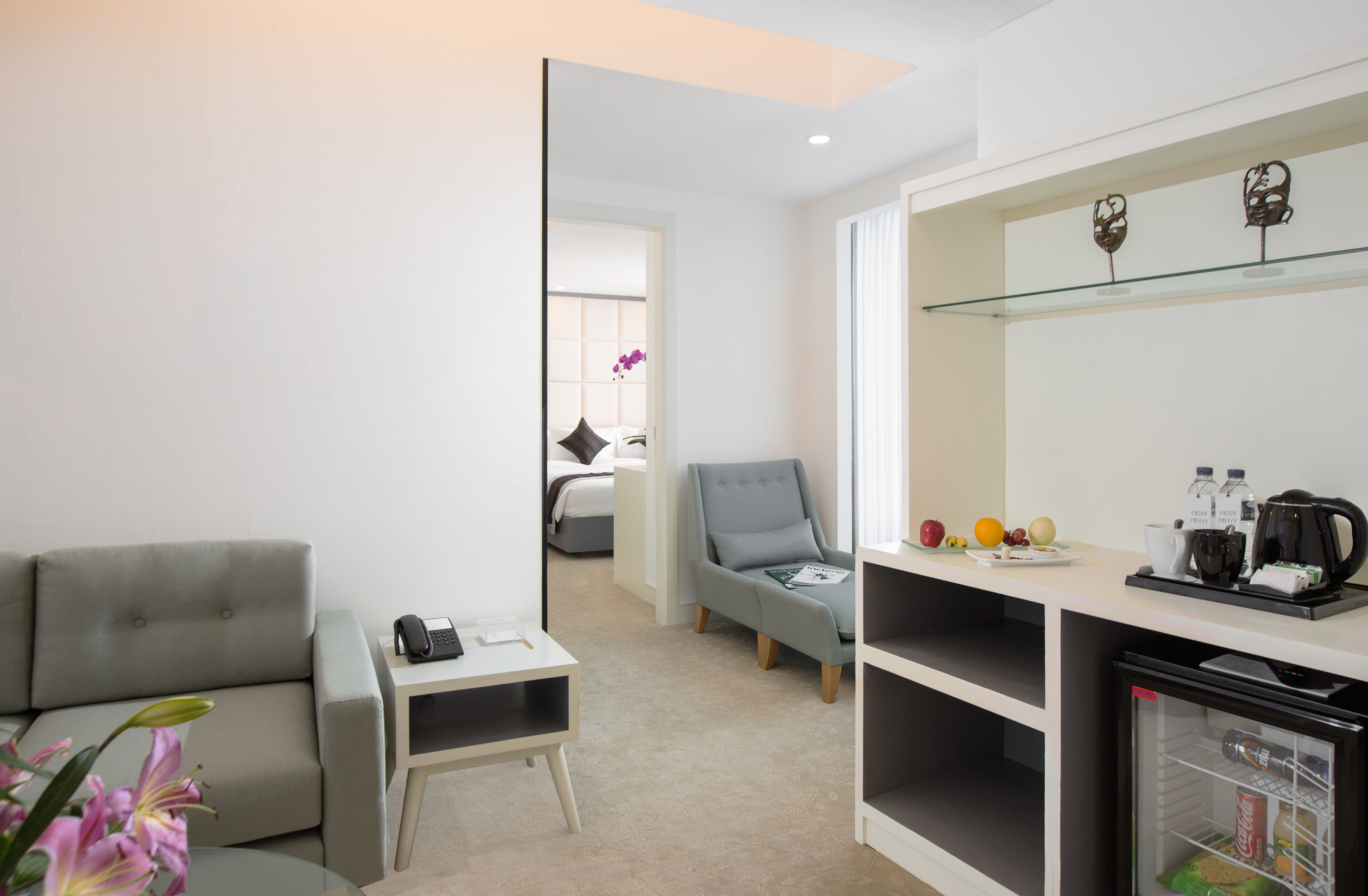 Modern hotel suite with grey sofa, armchair, mini-bar, and separate bedroom.