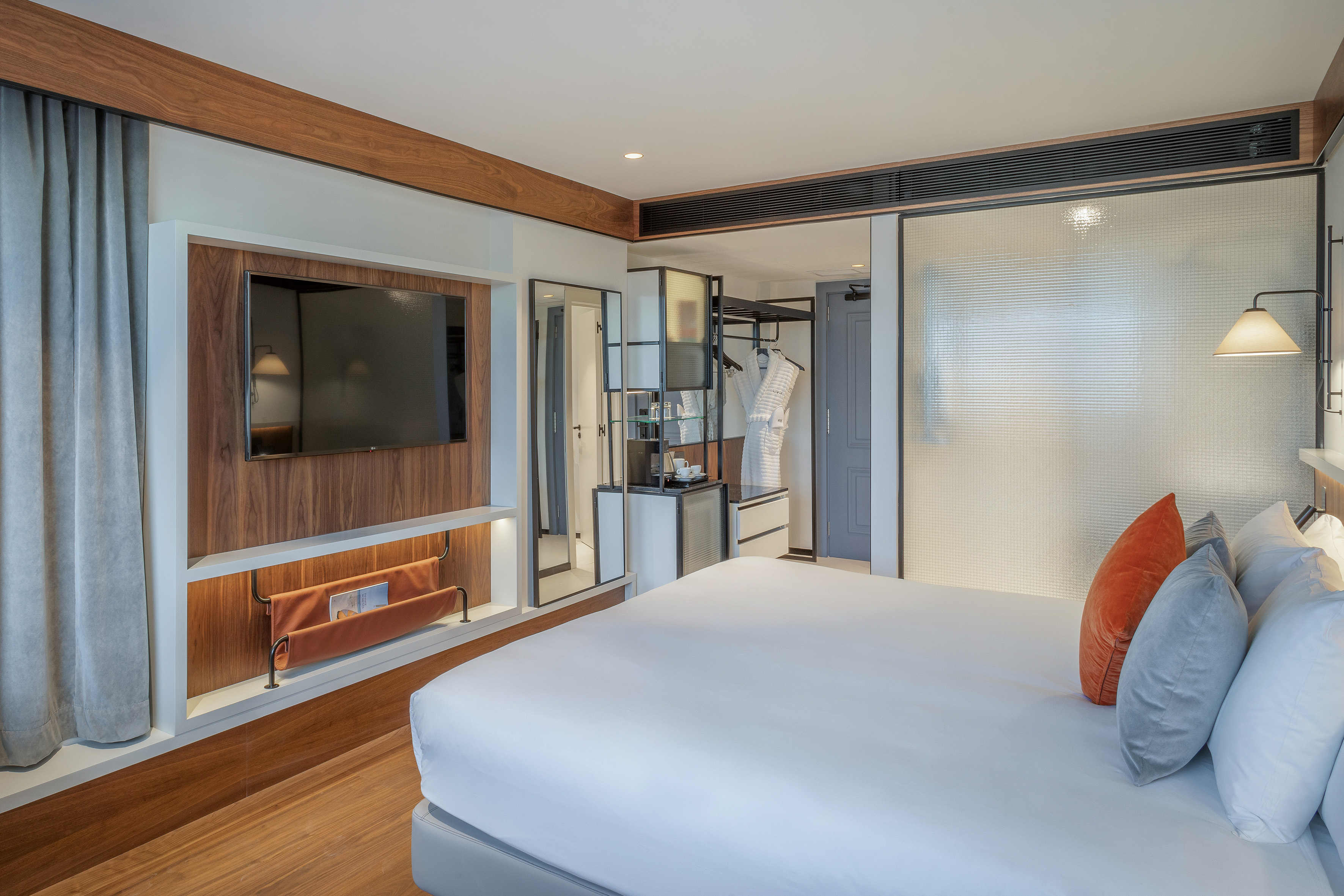 Modern hotel bedroom with bed, wall TV, wooden decor, and open closet area.
