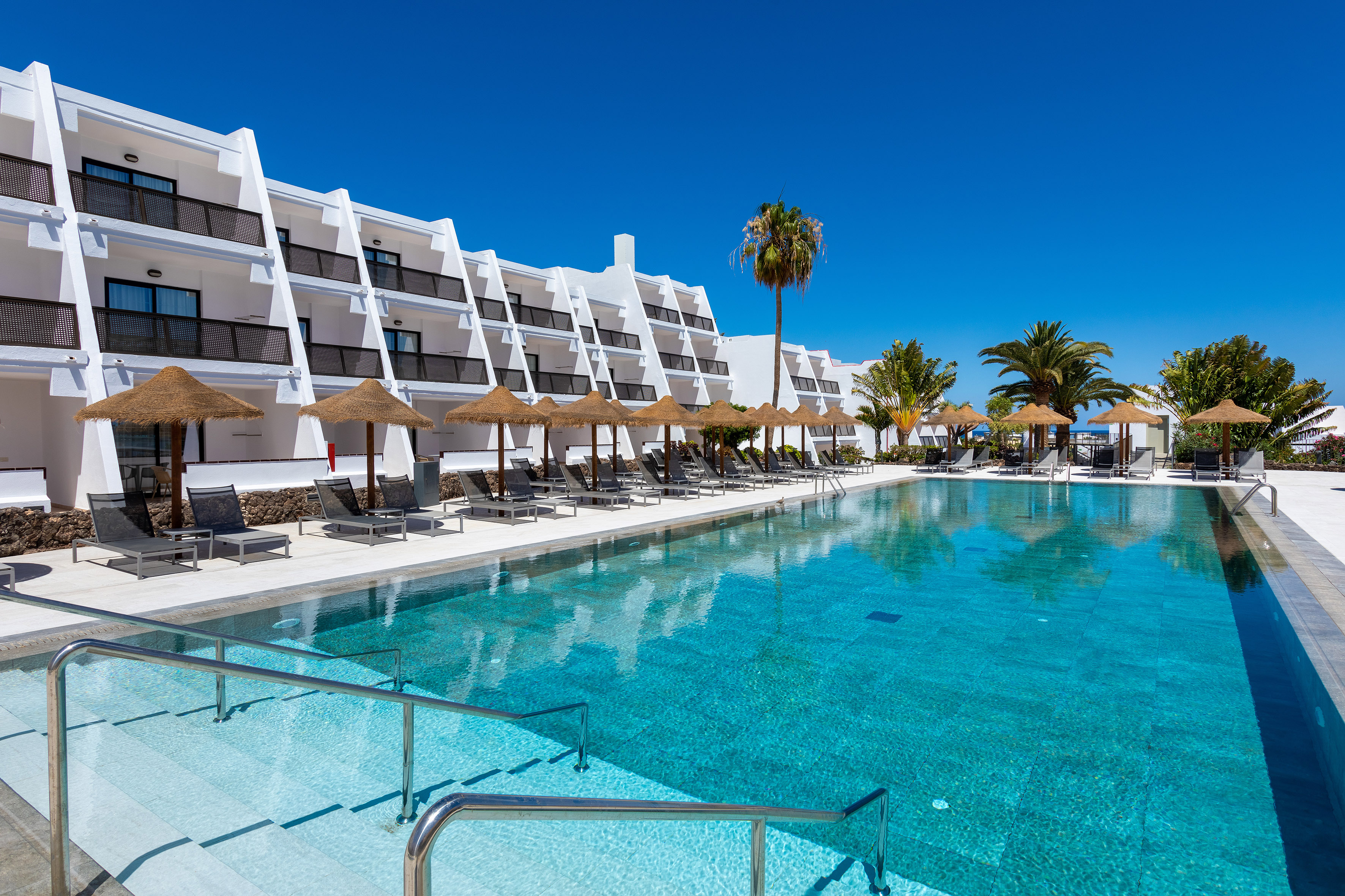 Modern hotel with large blue swimming pool, sun loungers, and palm trees.