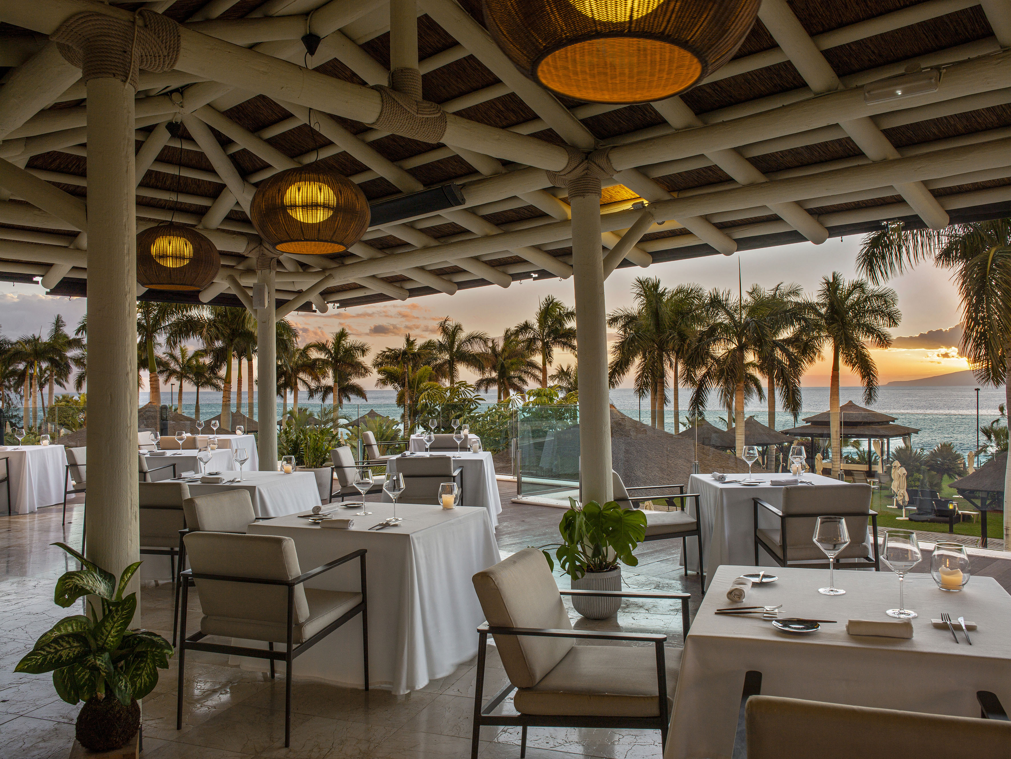 Elegant tropical outdoor restaurant overlooking ocean, palm trees, and sunset.