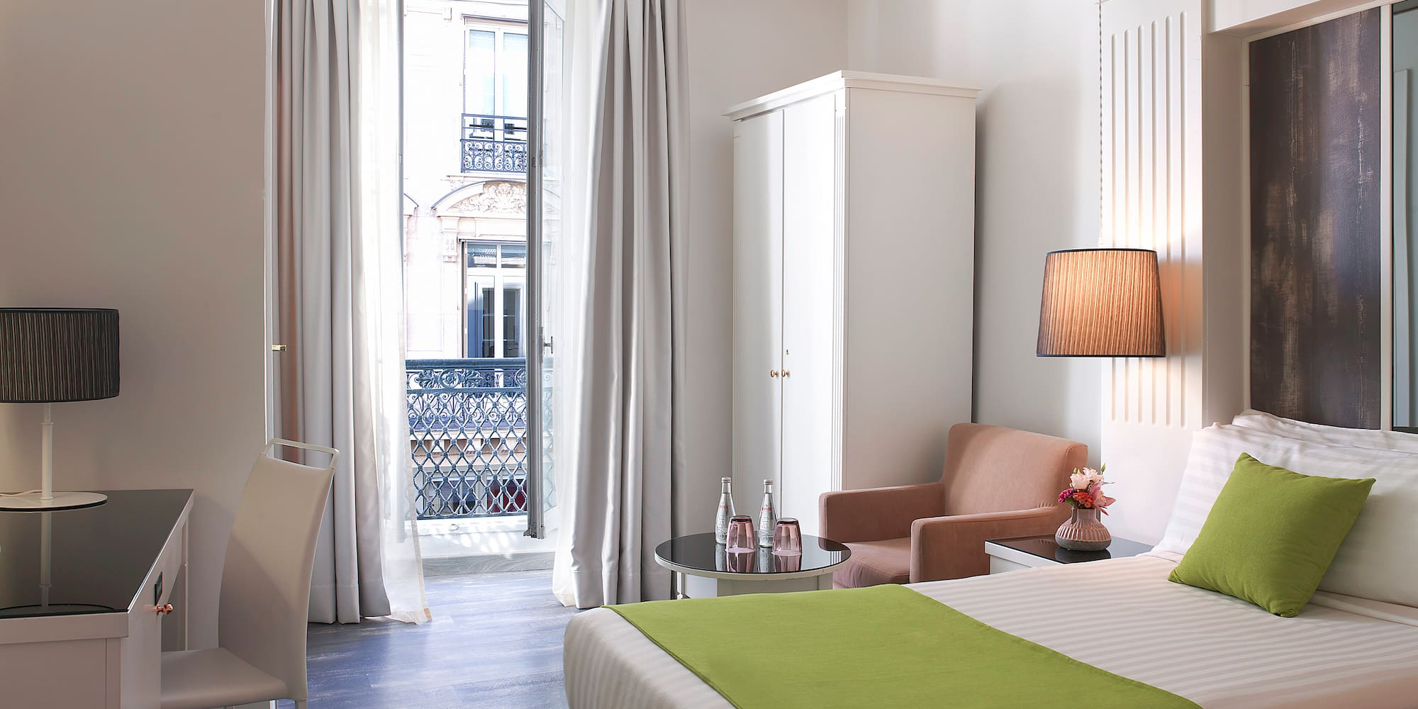 Hotel room with twin beds, green accents, desk, and balcony city view.