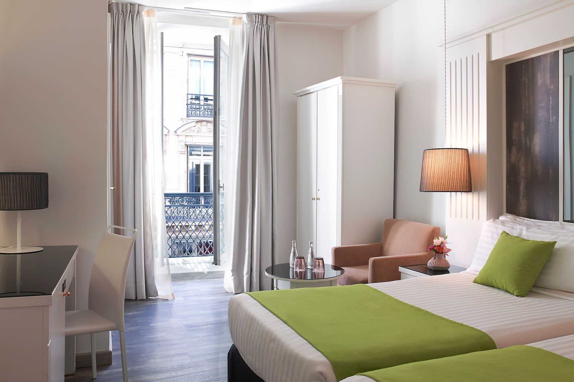 Hotel room with twin beds, green accents, desk, and balcony city view.
