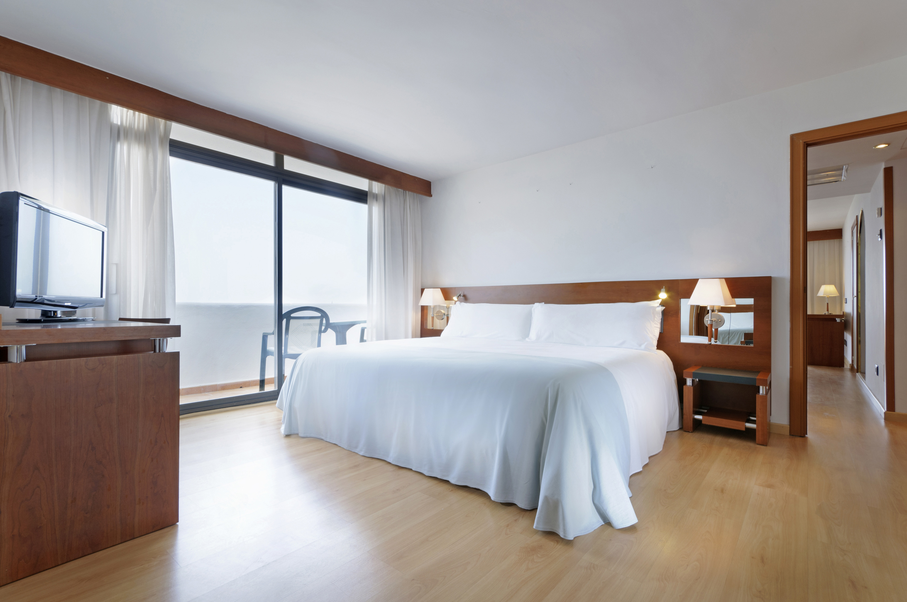 Modern hotel bedroom with king bed, balcony access, wooden floors, and TV.