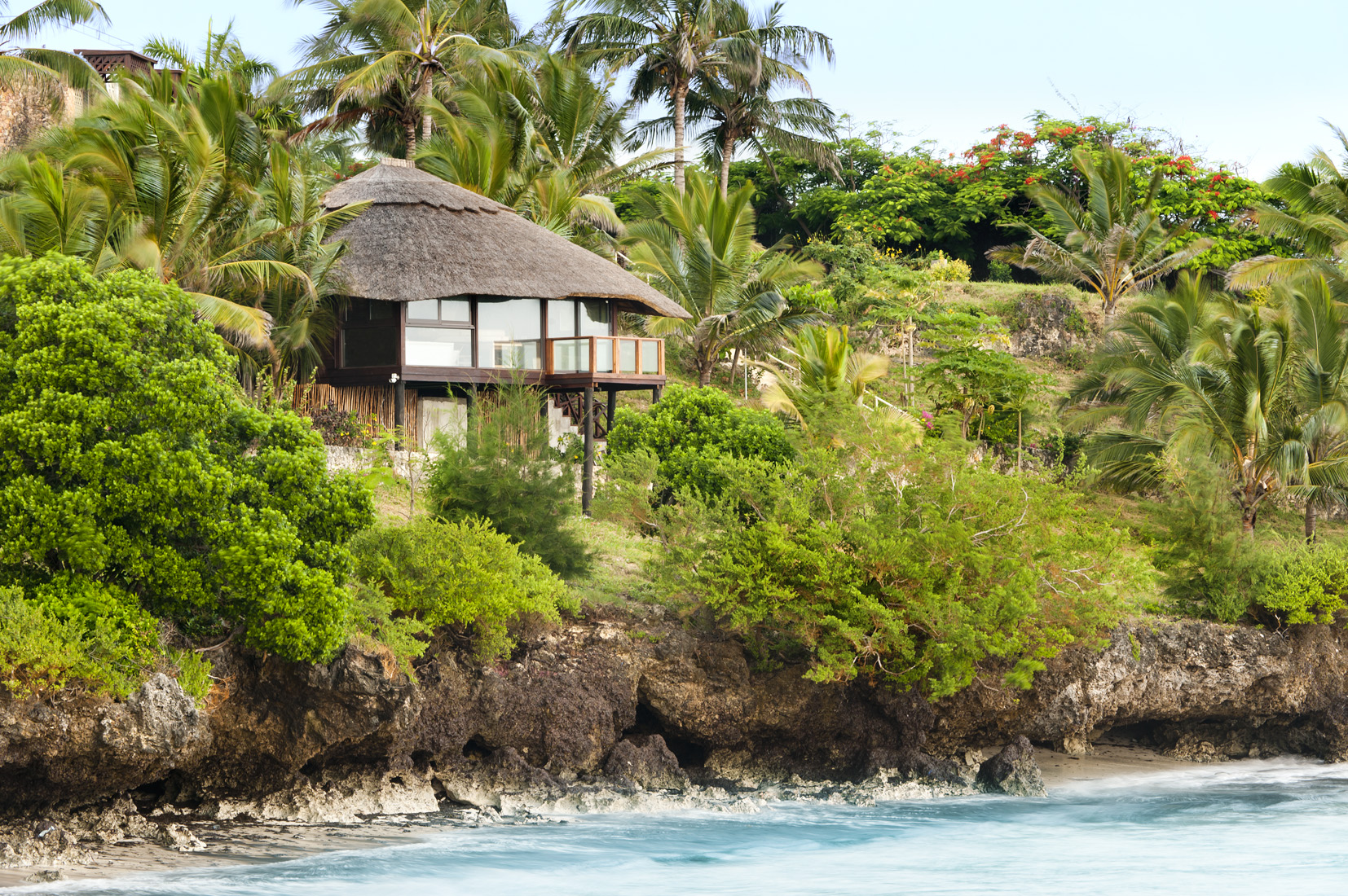 Tropical cliffside bungalow with thatched roof, palm trees, and ocean view.