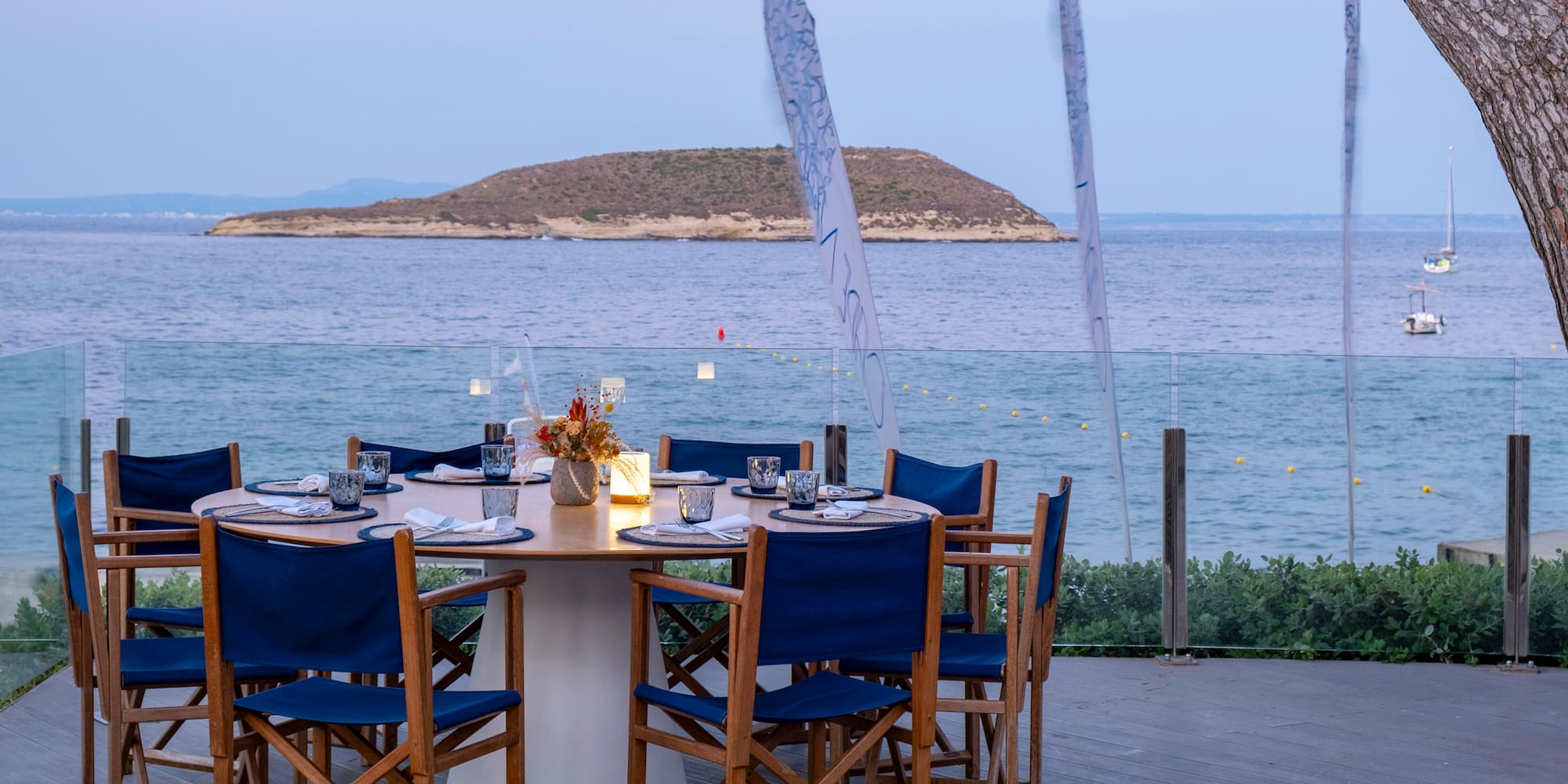 a table set up with blue chairs and a tree overlooking the water