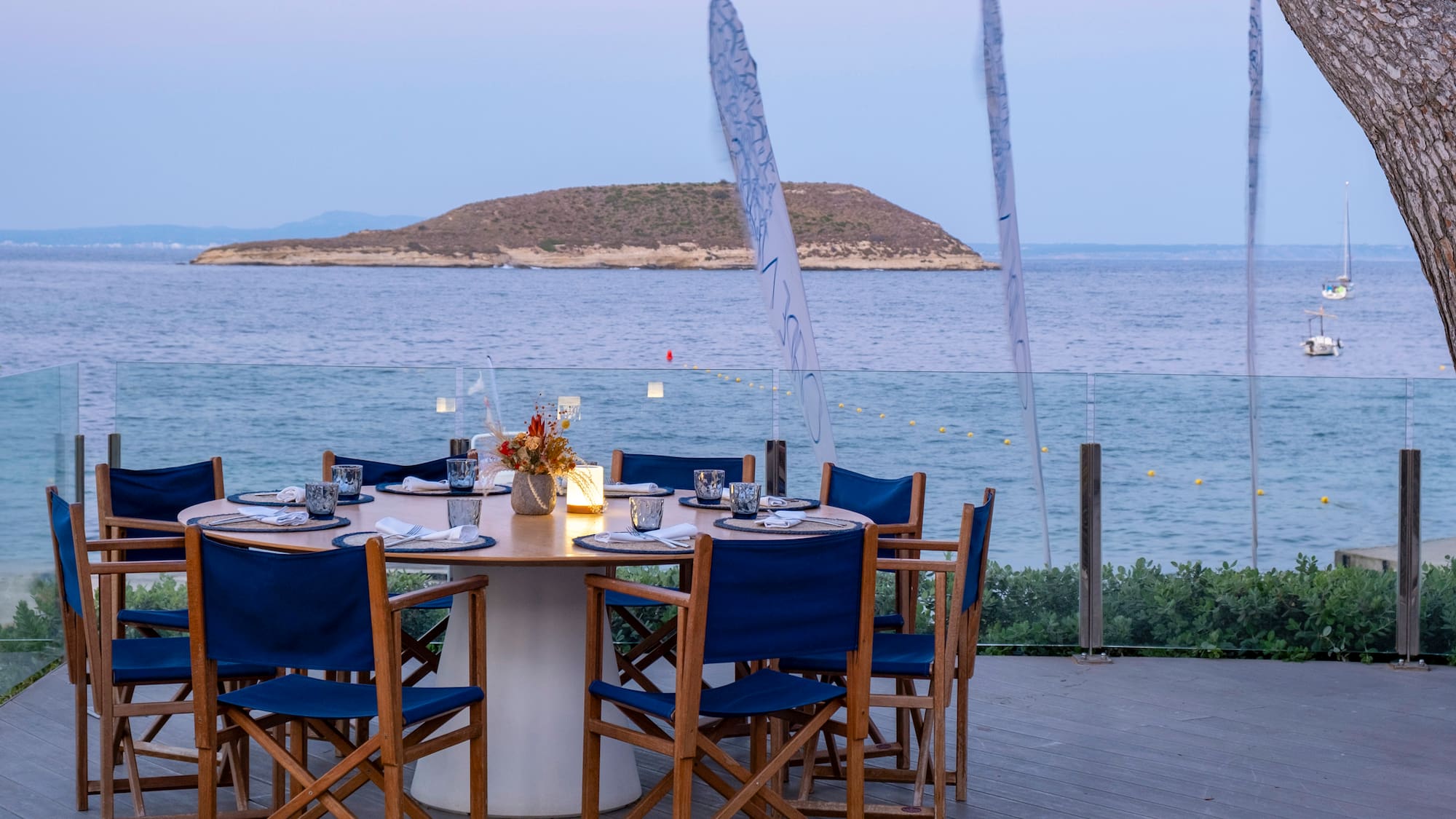 a table set up with blue chairs and a tree overlooking the water