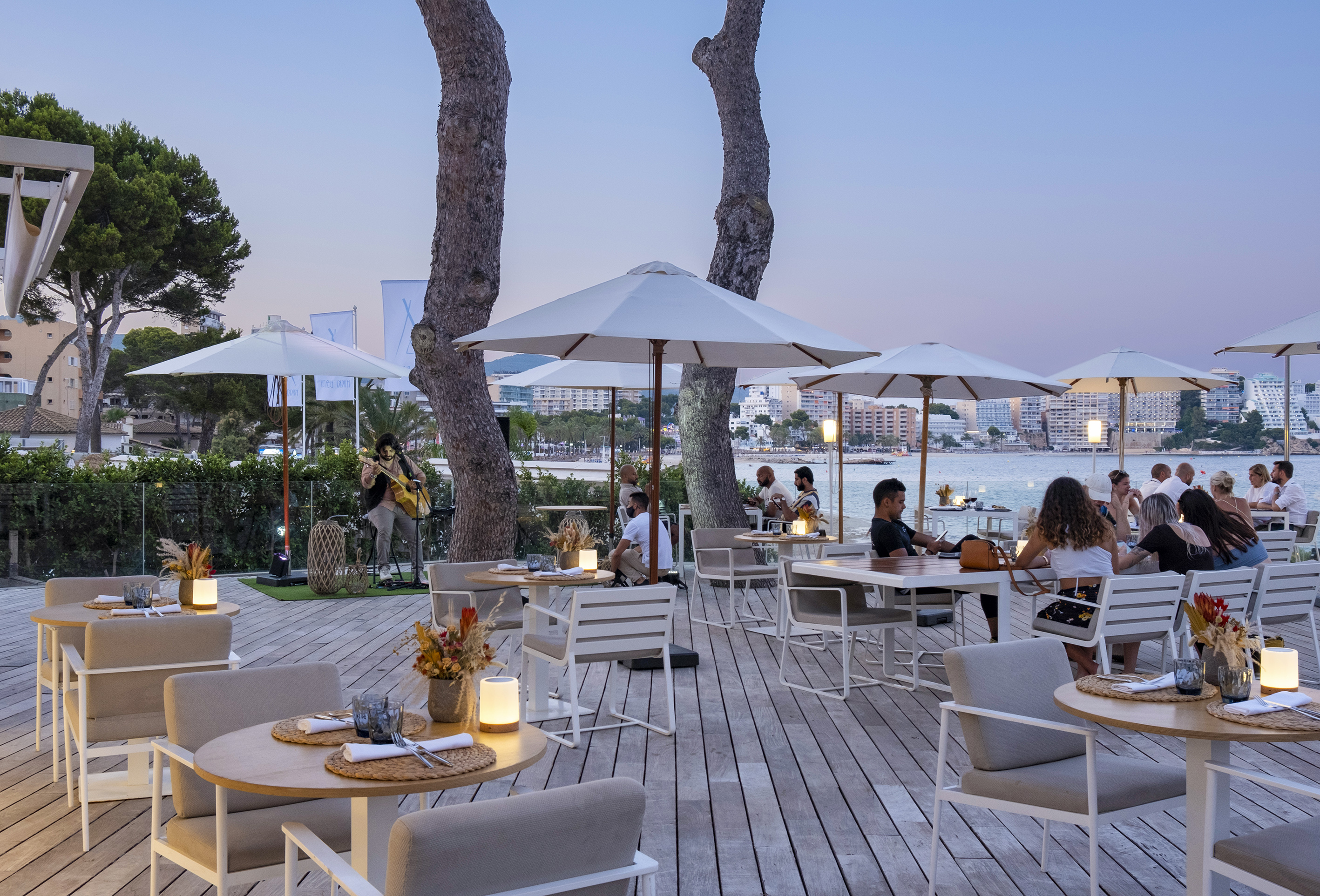 Beachfront restaurant terrace with diners, live music, and city view at dusk.