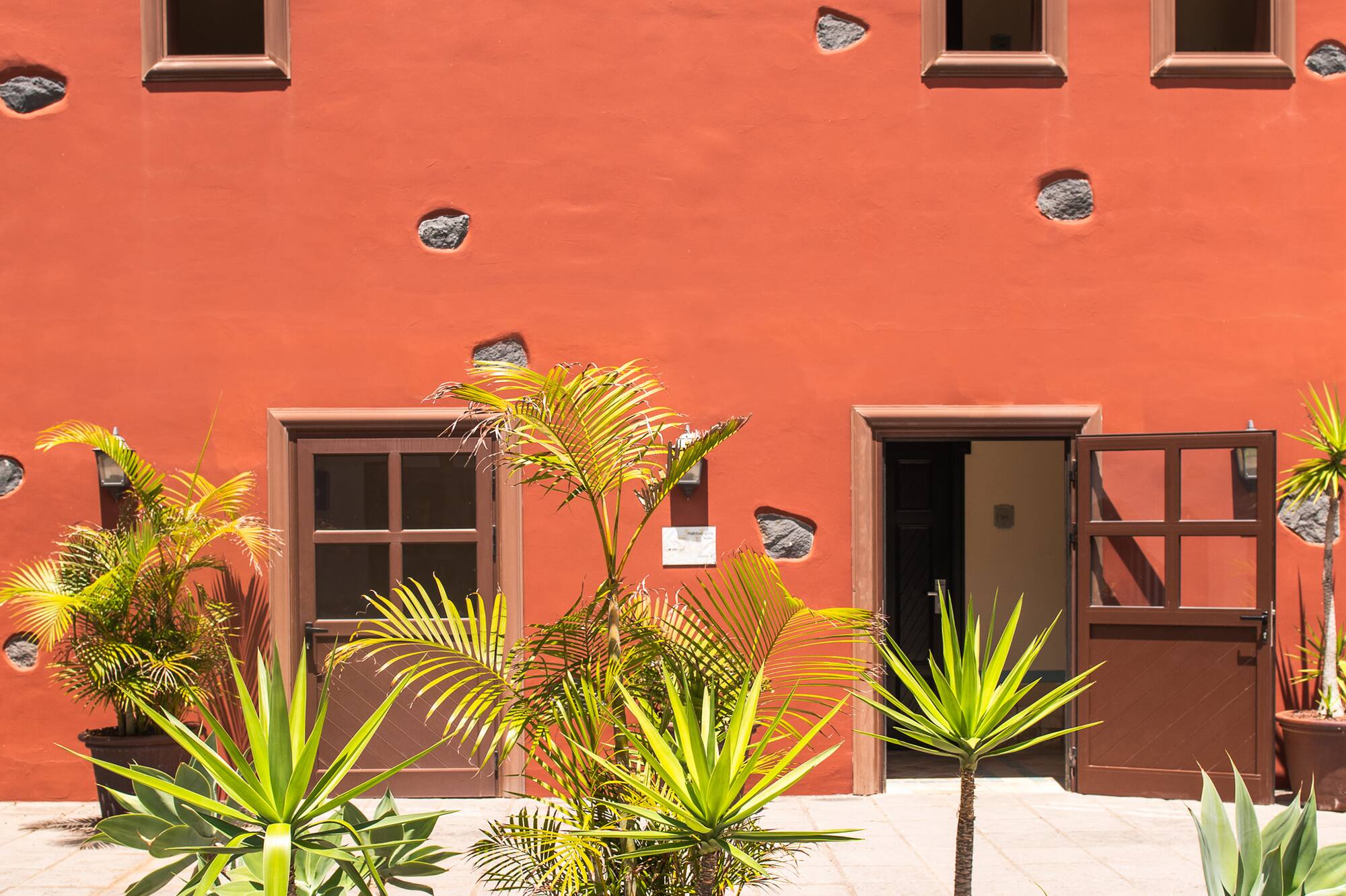 Red building with terracotta roof, potted tropical plants in a sunny courtyard.