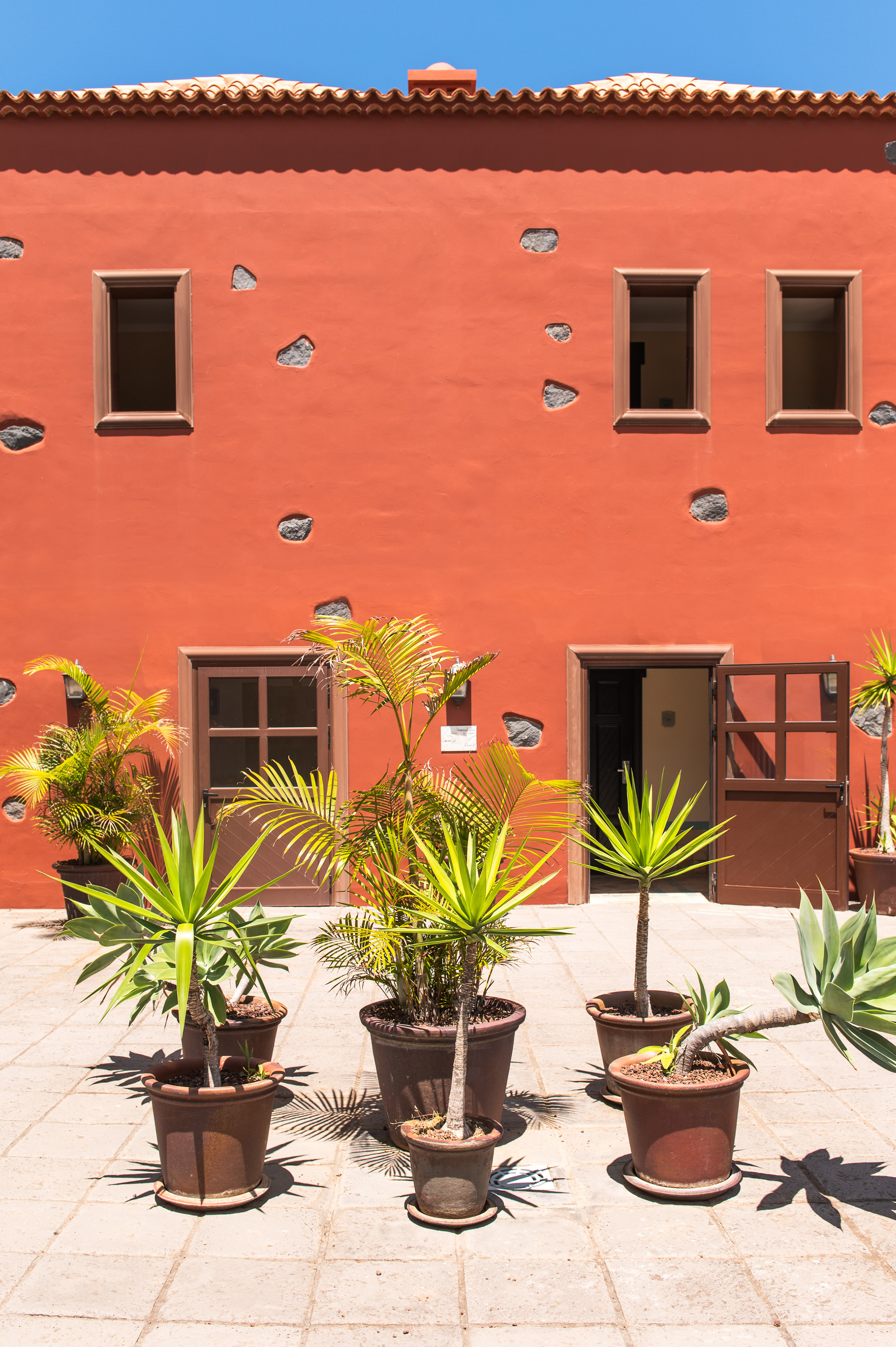 Red building with terracotta roof, potted tropical plants in a sunny courtyard.