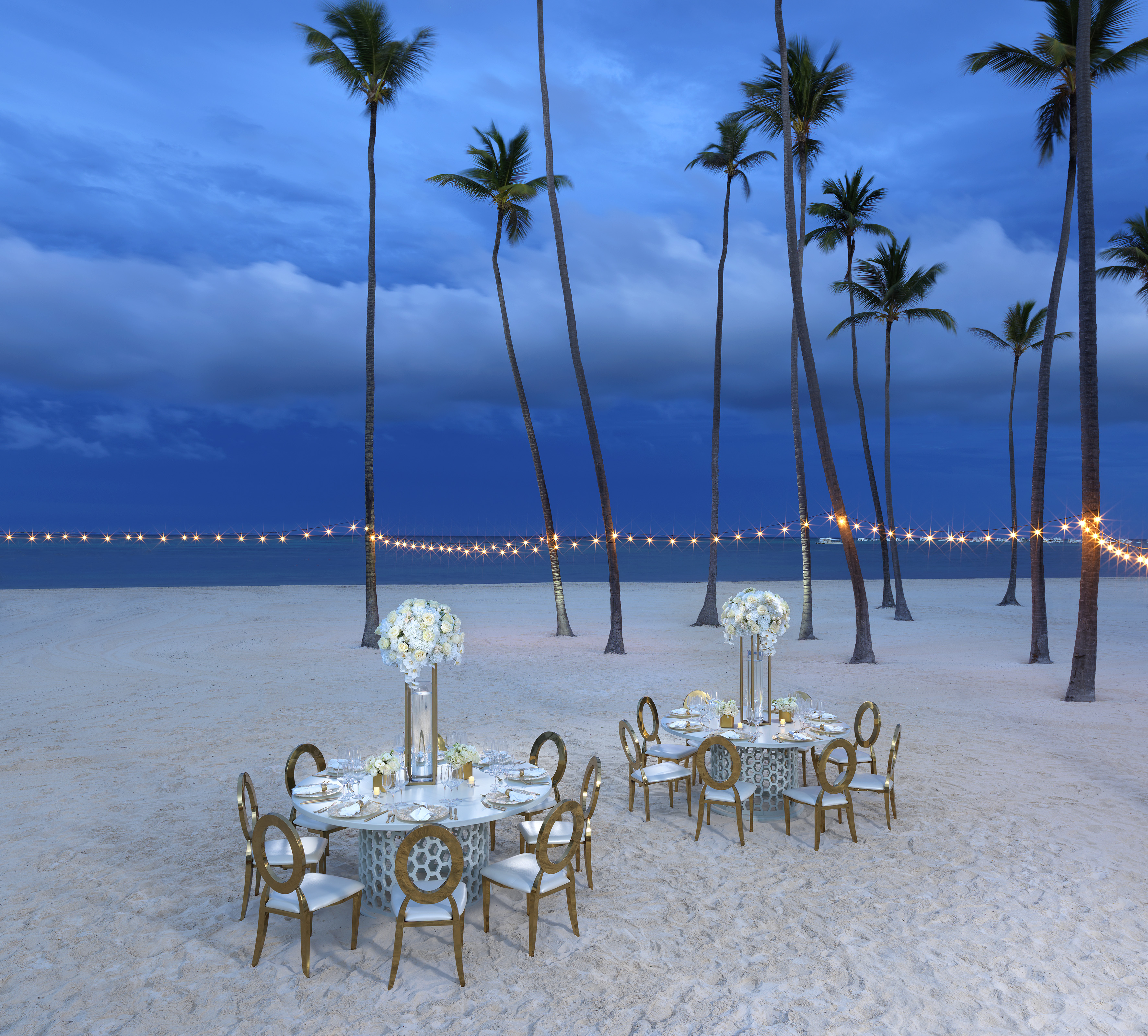Beach dining with gold chairs, floral decor, palm trees, string lights at night.
