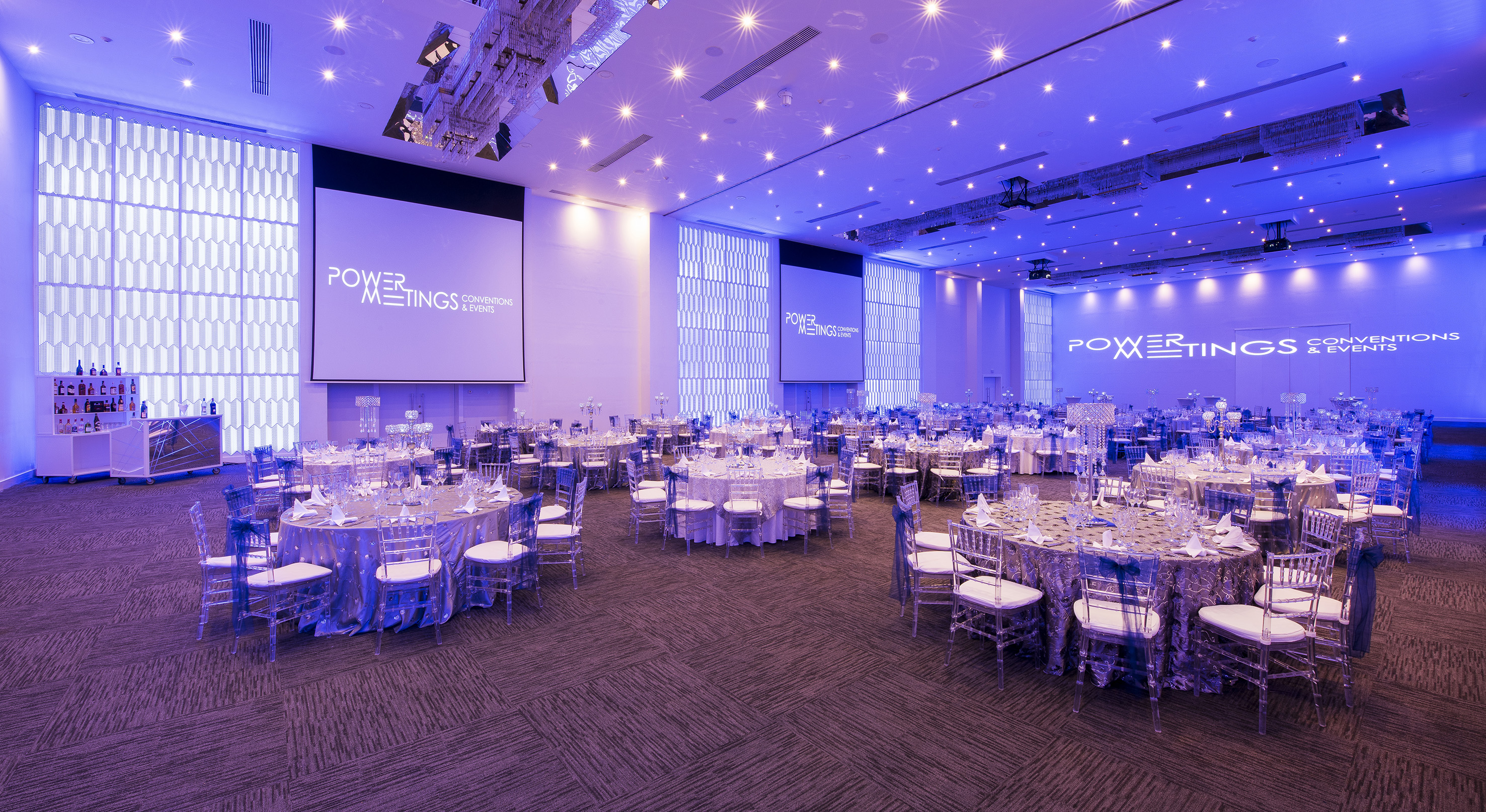 Elegant event hall with banquet tables, clear chairs, and blue lighting.