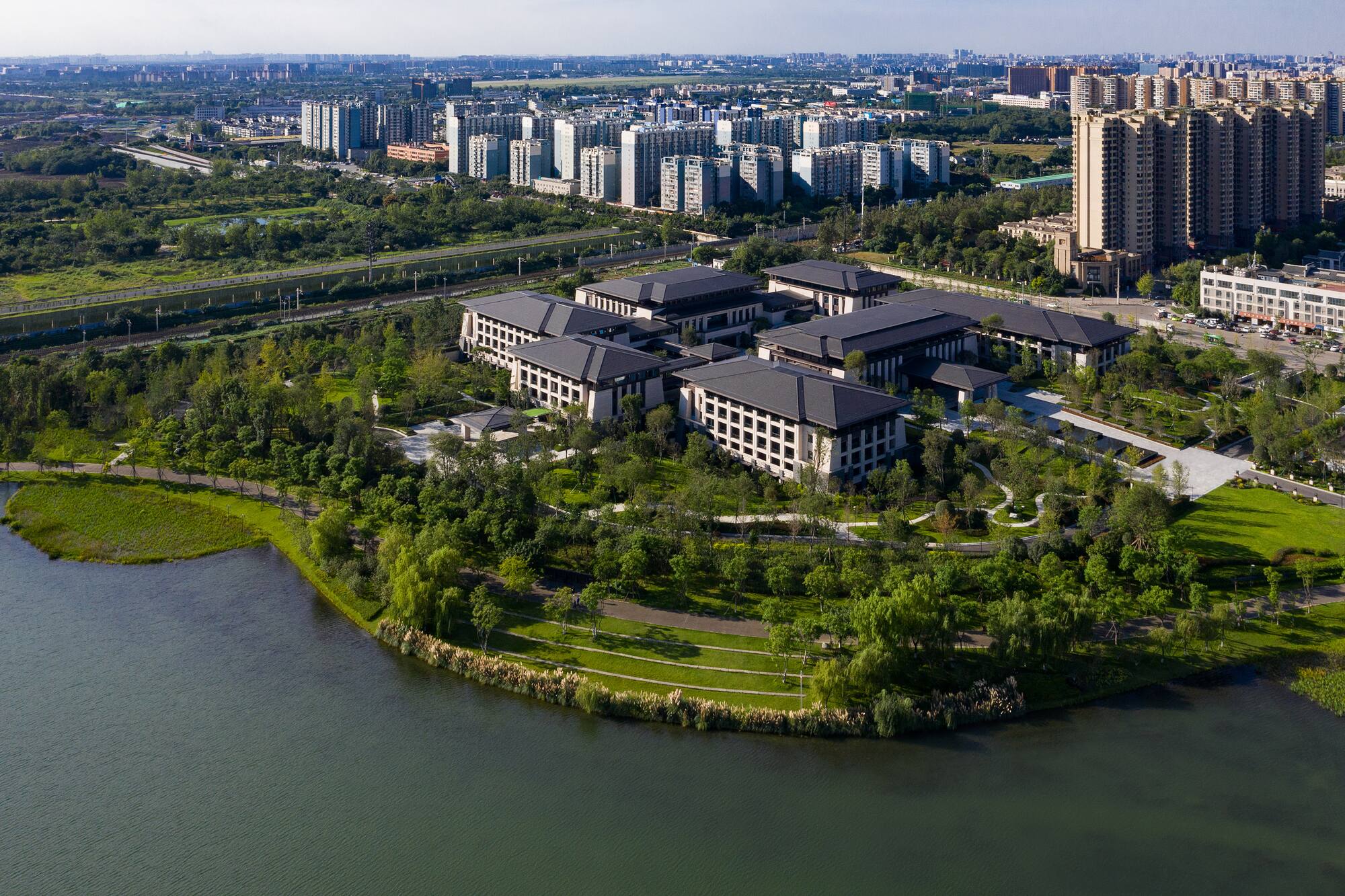Aerial view: Lakeside campus with modern buildings, lush gardens, and urban skyline.