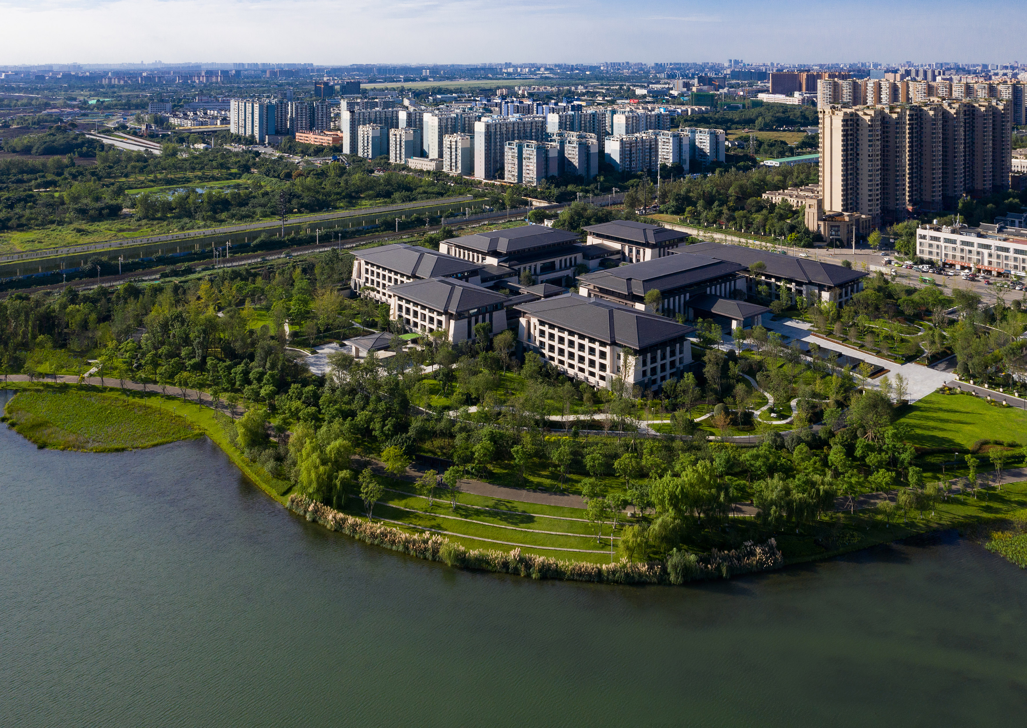 Aerial view: Lakeside campus with modern buildings, lush gardens, and urban skyline.