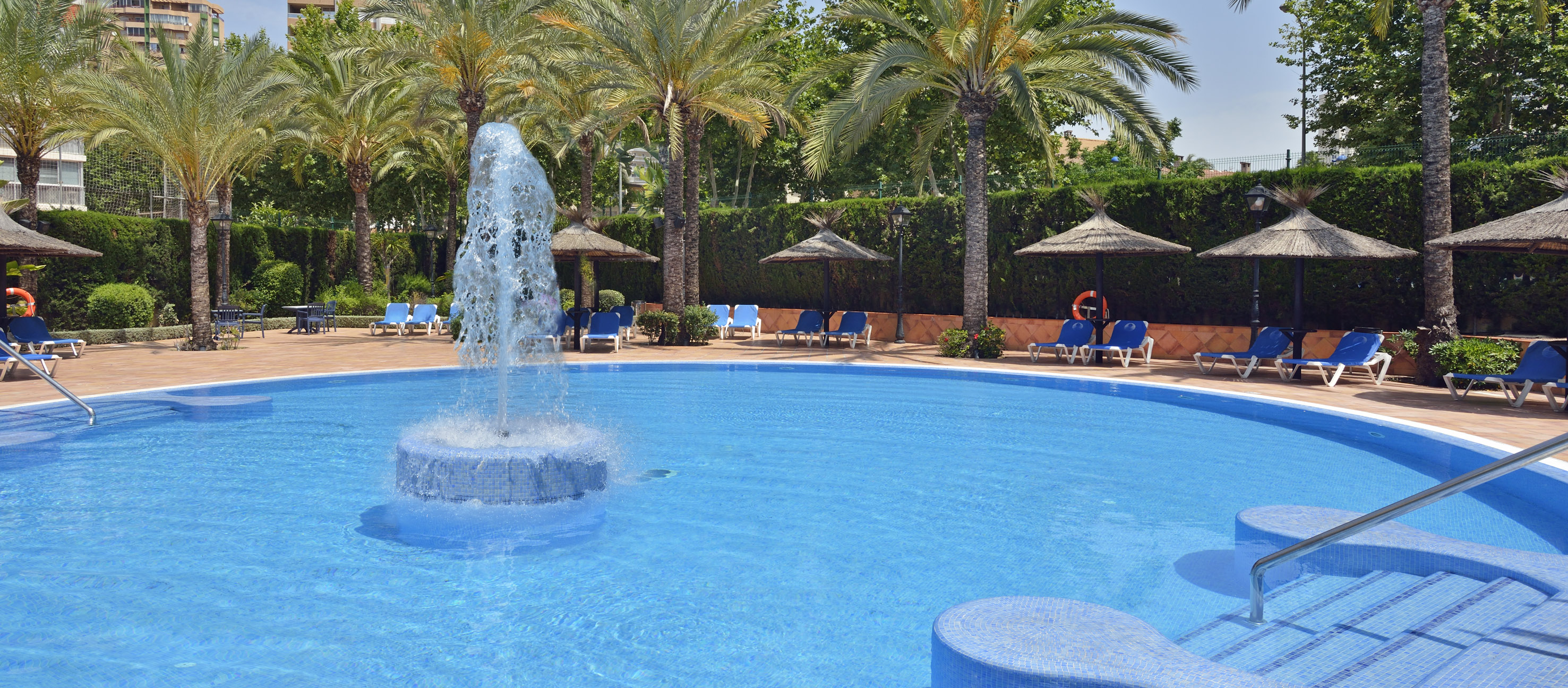 Resort swimming pool with fountain, palm trees, and sun loungers.