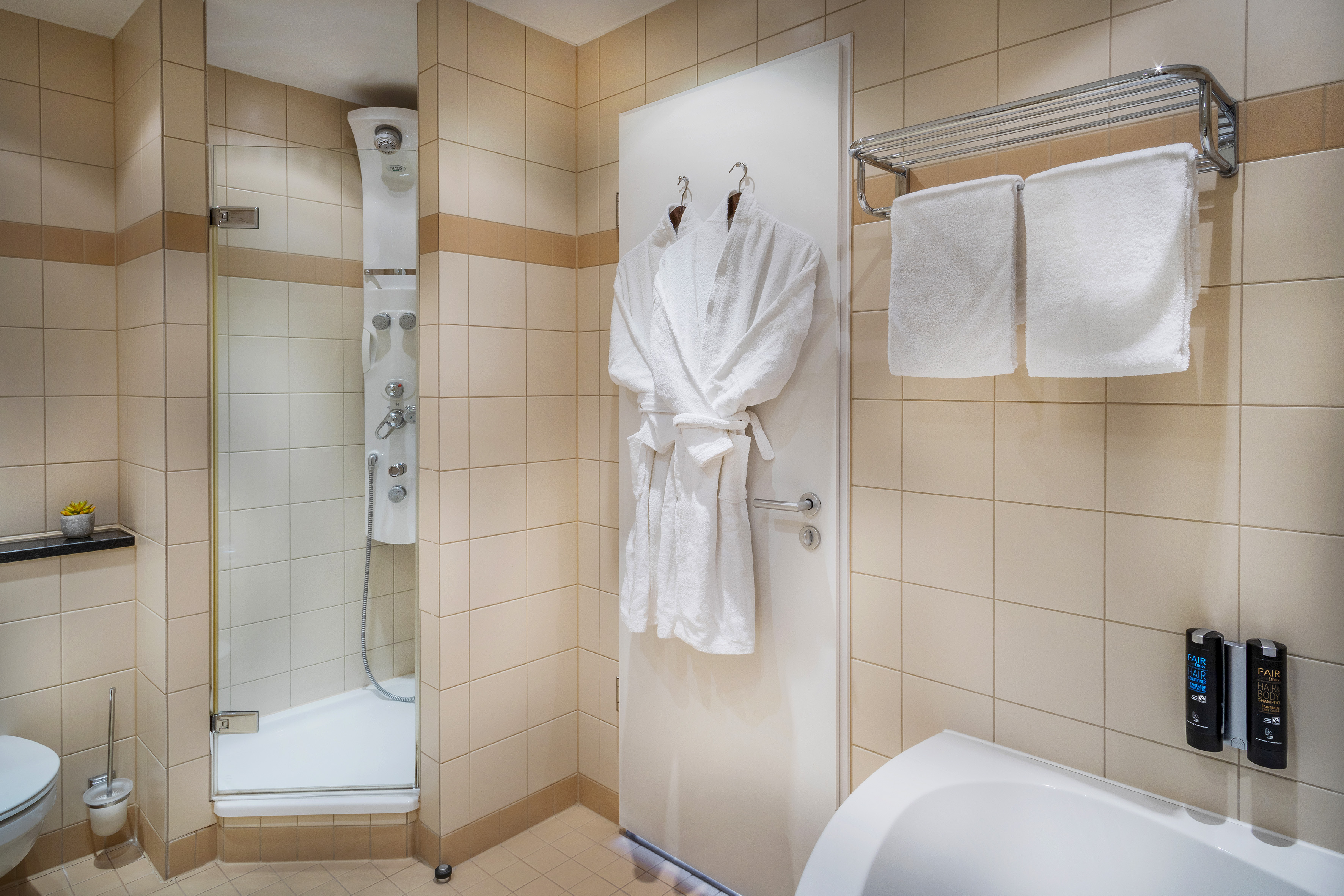 Modern tiled bathroom with multi-jet shower, bathtub, bathrobes, and fresh towels.