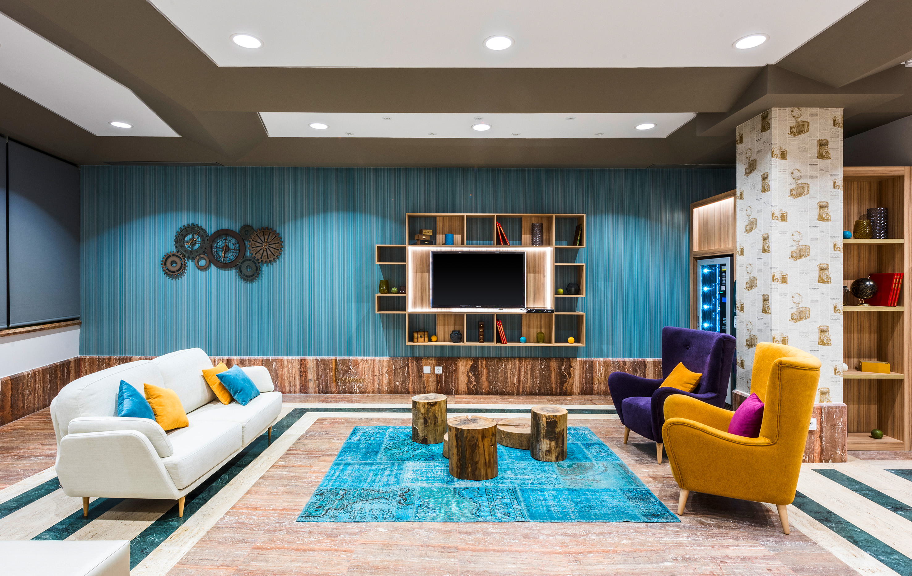 Modern lounge: blue wall, white sofa, colorful chairs, wood tables, and TV unit.