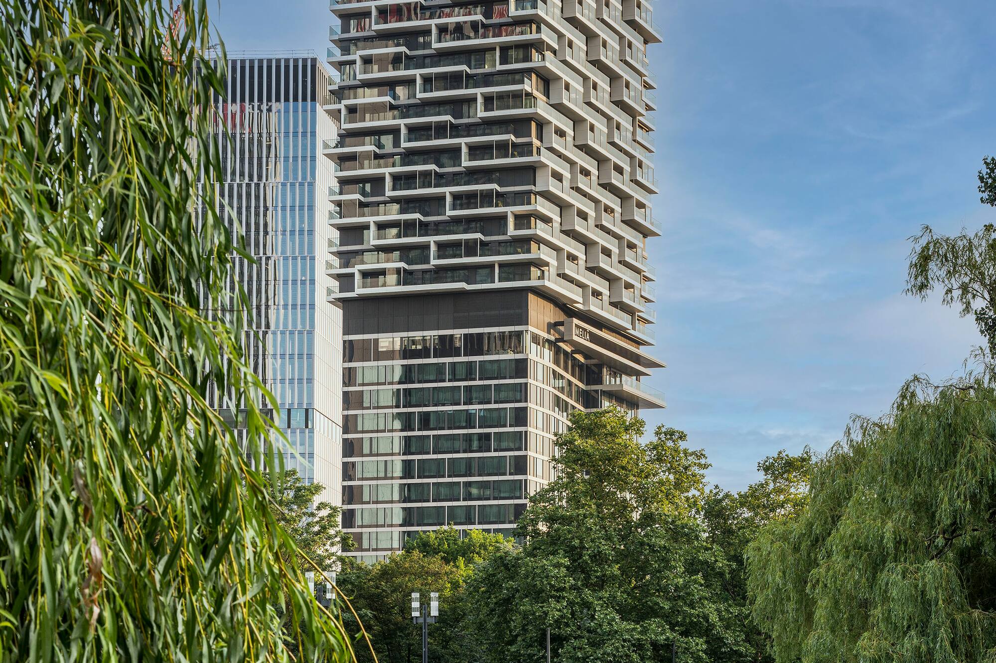 Modern Meliá Hotel with staggered balconies by a park lake and willow tree.