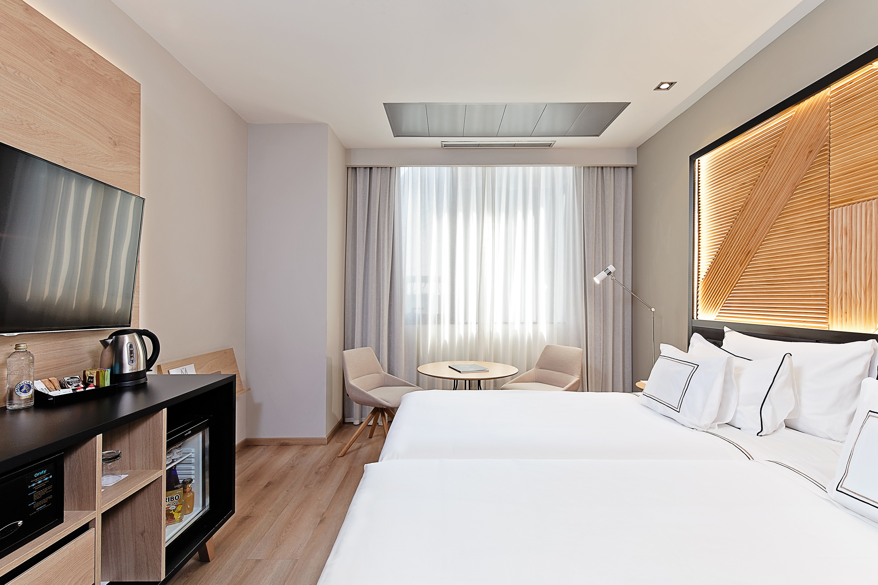 Modern hotel room with twin beds, wooden headboard, TV, and mini-bar.