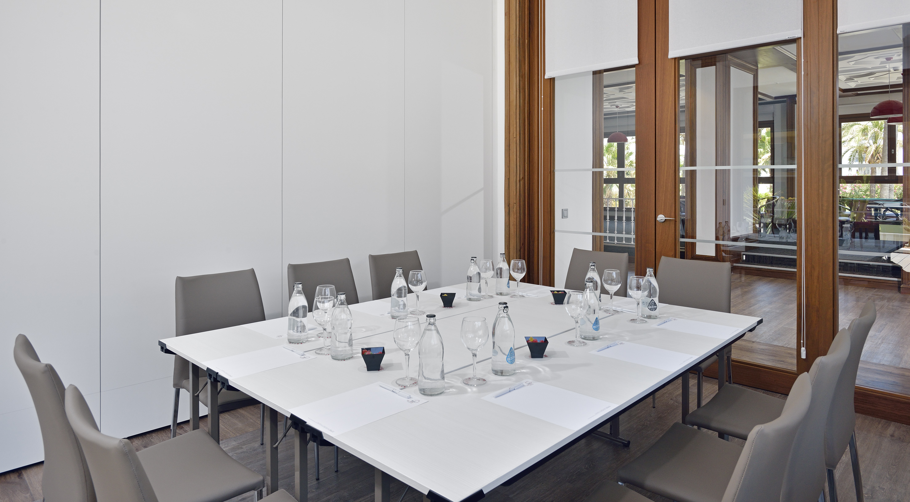 Conference room with white table, grey chairs, water bottles, and glass doors.