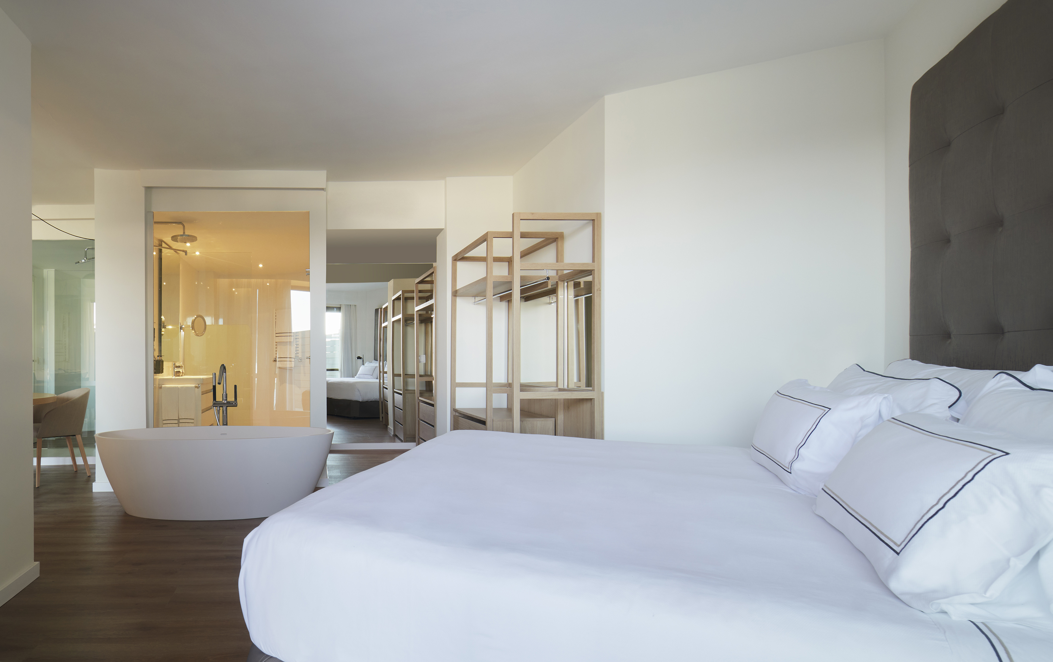 Modern hotel suite with white bed, freestanding bathtub, and open wardrobe.