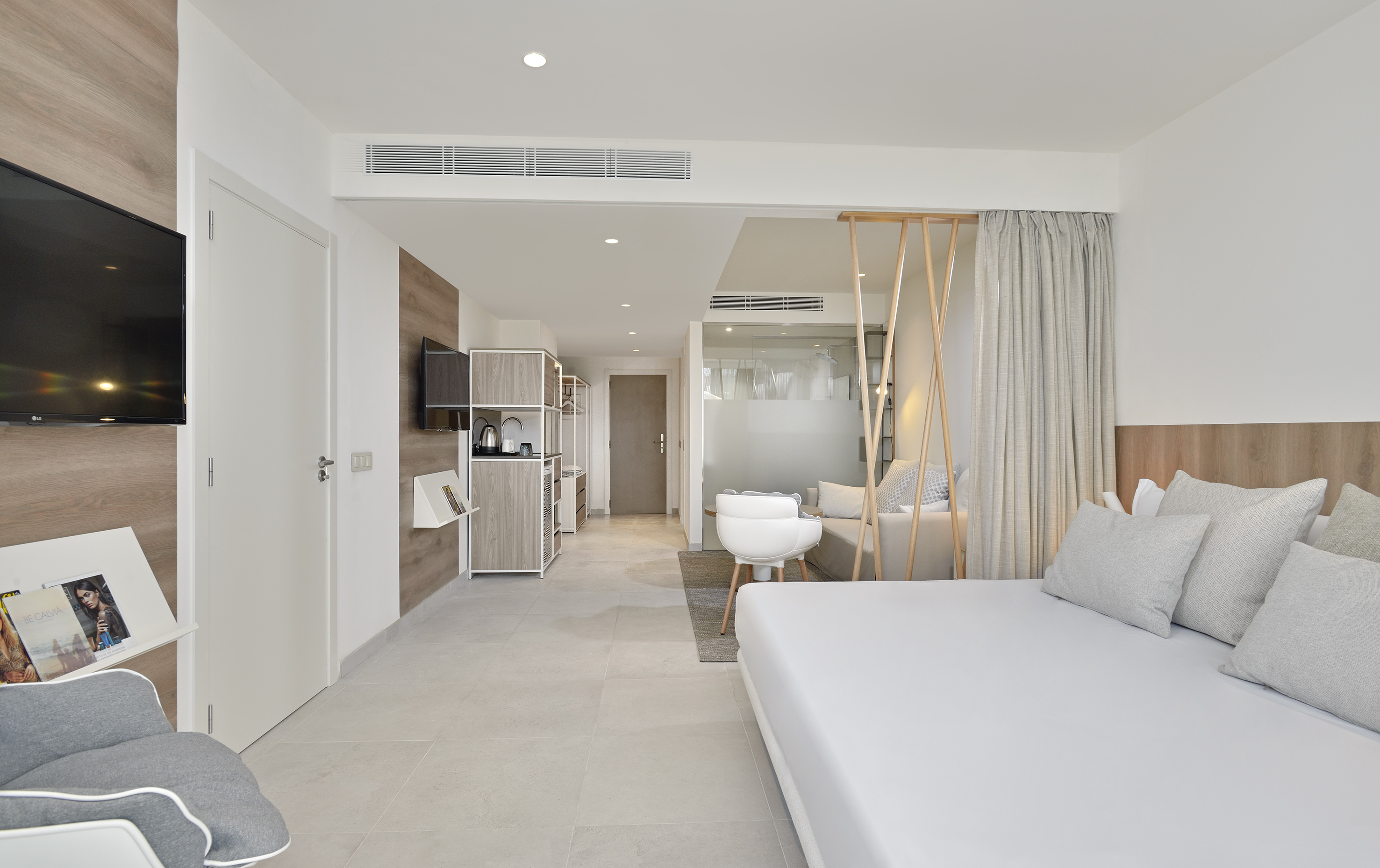 Spacious modern hotel room with white bed, sofa, TV, and light wood decor.