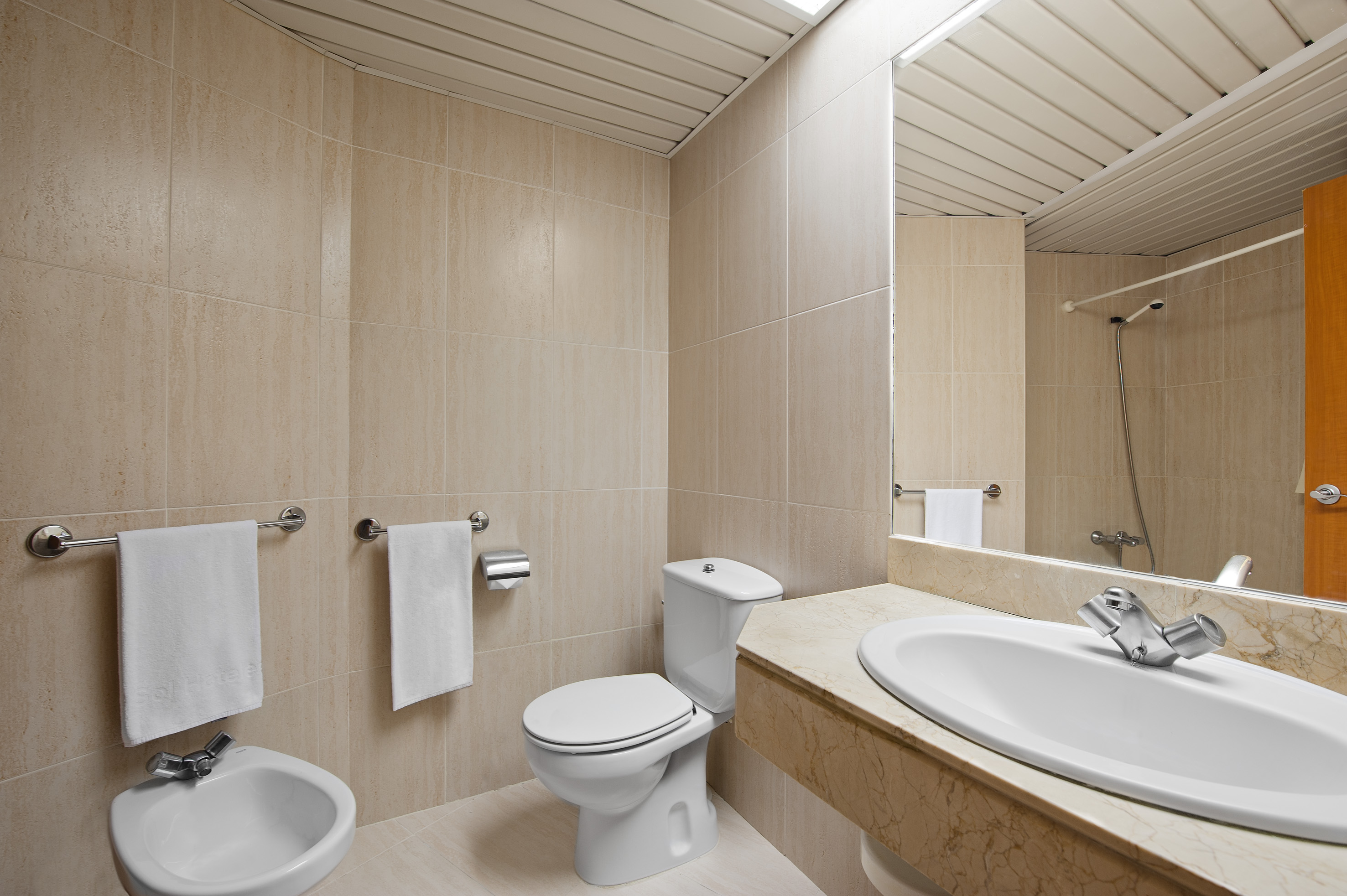 Modern hotel bathroom with beige tiled walls, white toilet, sink, and bidet.