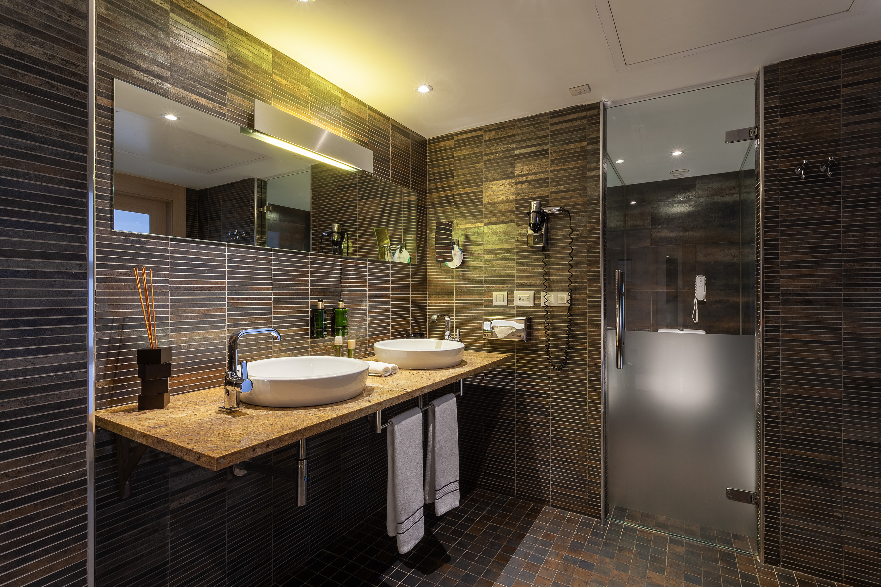 Modern bathroom with twin sinks, dark tiled walls, and a glass shower.