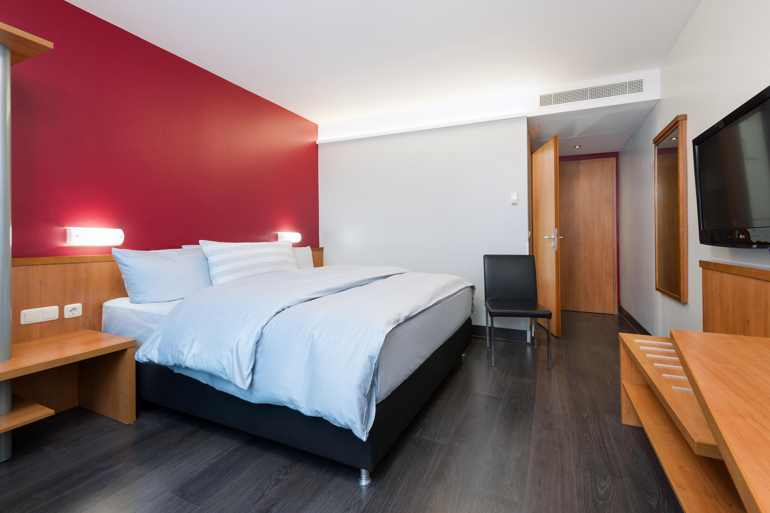 Modern hotel room with red accent wall, king bed, and dark wood floor.
