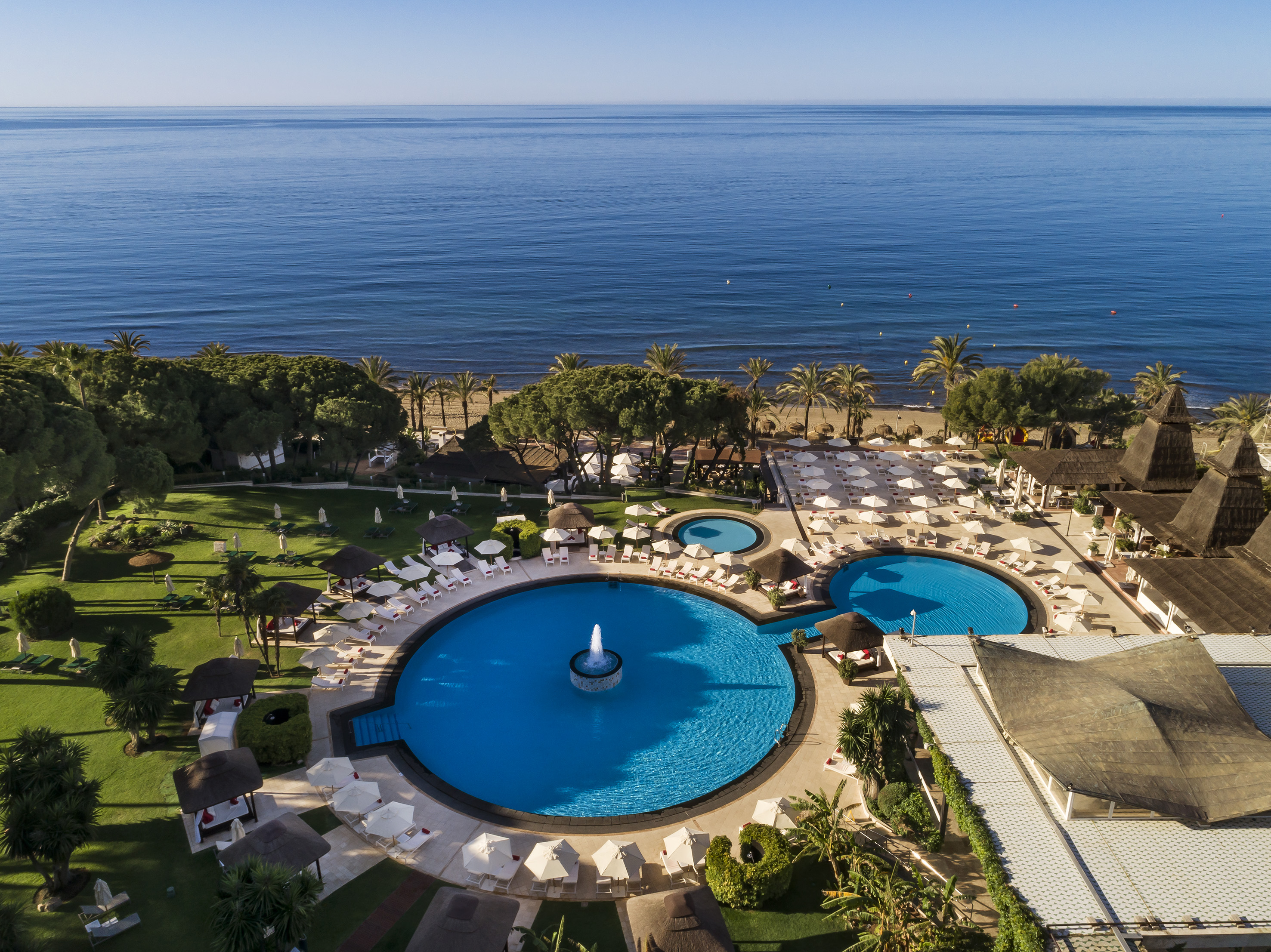 Aerial: Luxury resort beachfront pools, fountain, palm trees, ocean.