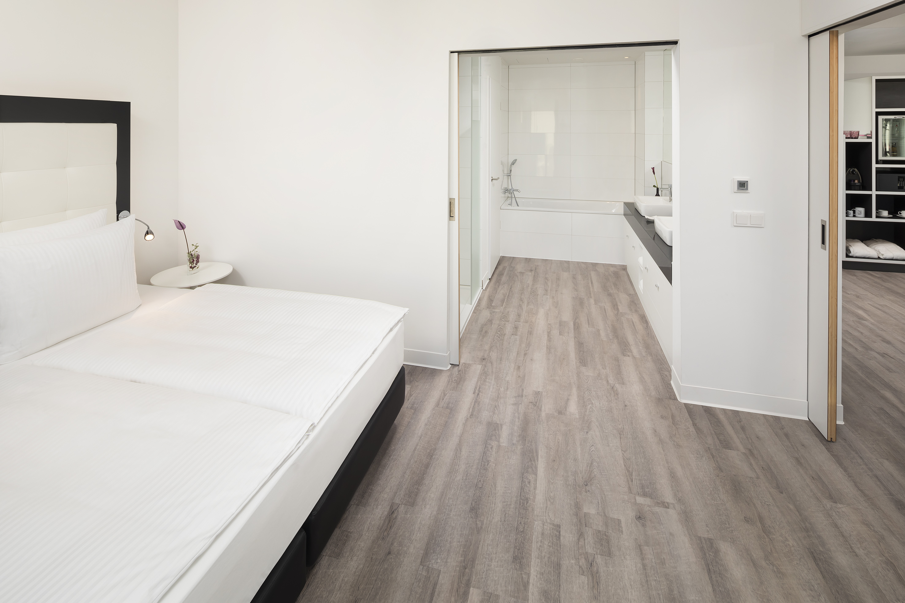 Modern hotel suite bedroom with white bed, wood floor, and open bathroom.
