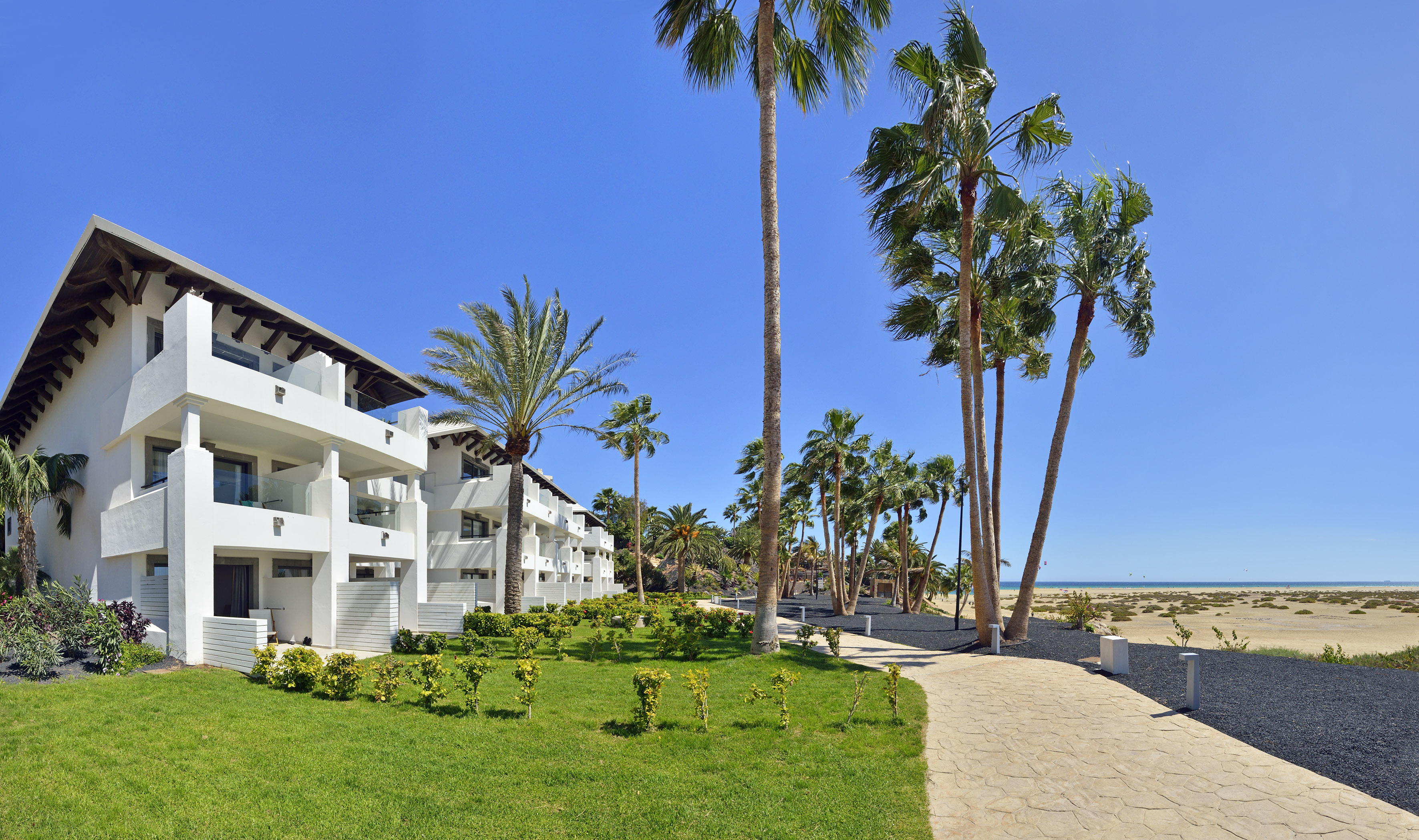 White beachfront resort with palm trees, green grass, and paved path to the ocean.