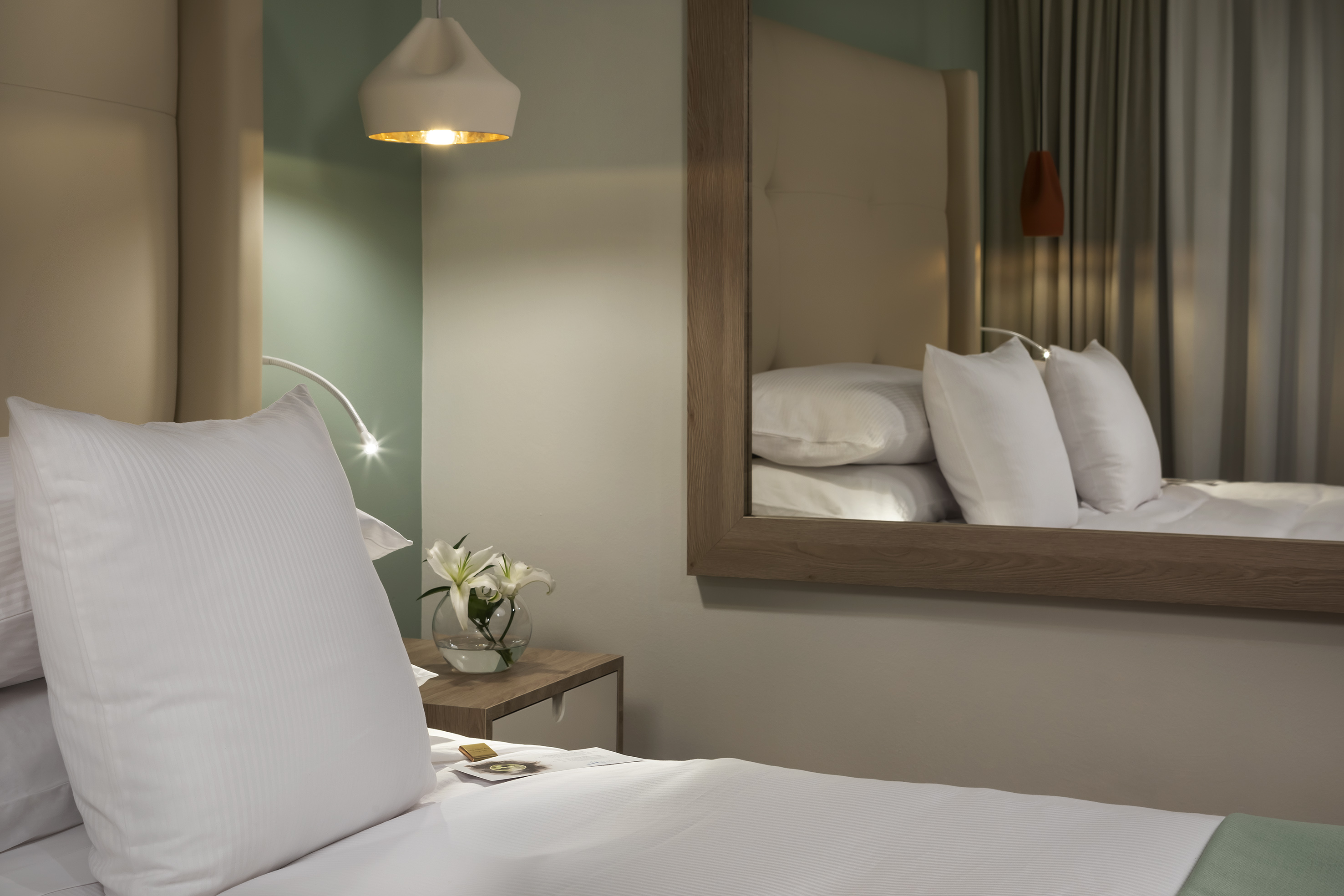 Modern hotel bedroom with white bed, nightstand, lilies, and reflective mirror.