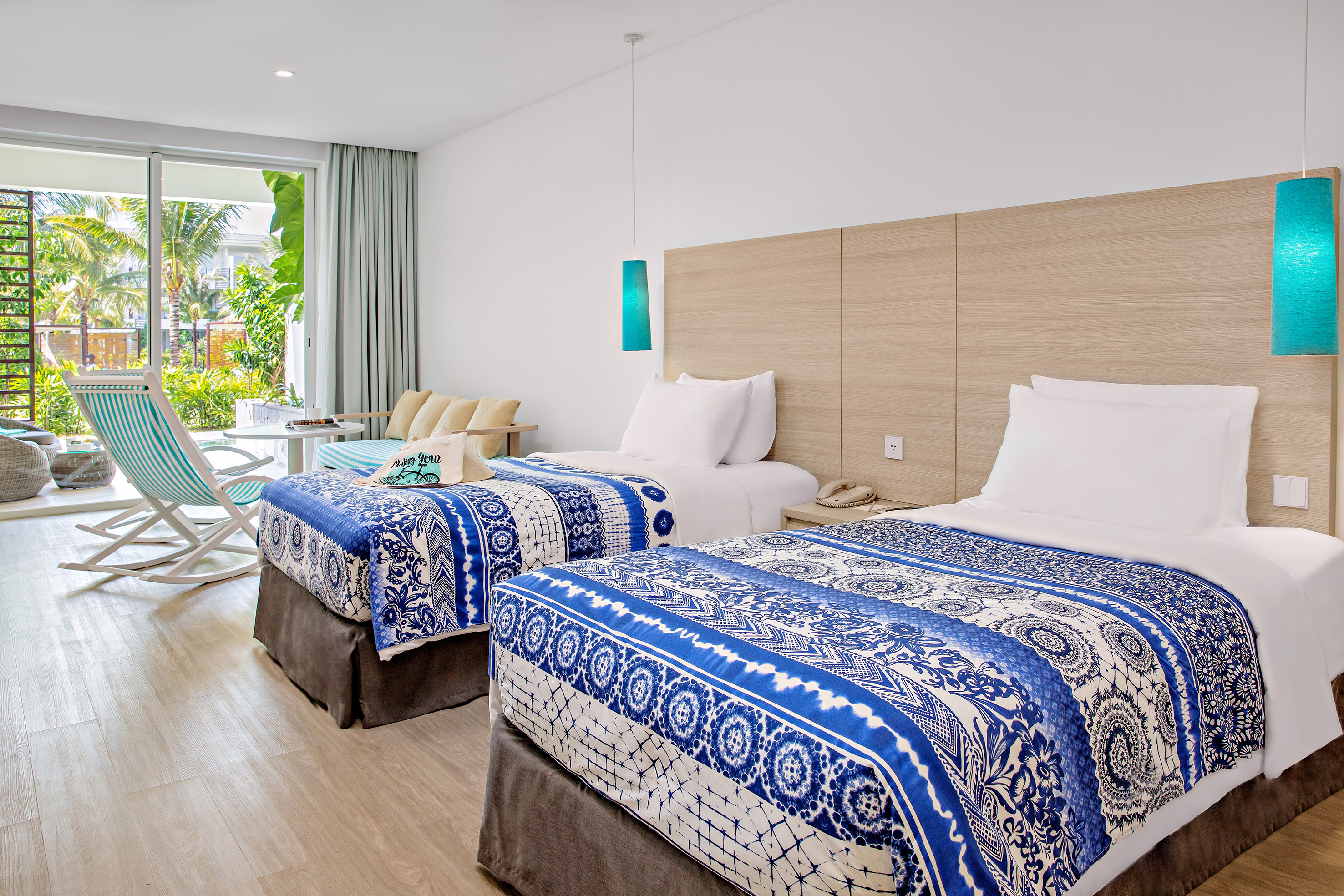 Modern hotel room with two twin beds, blue bedspreads, and tropical view balcony.