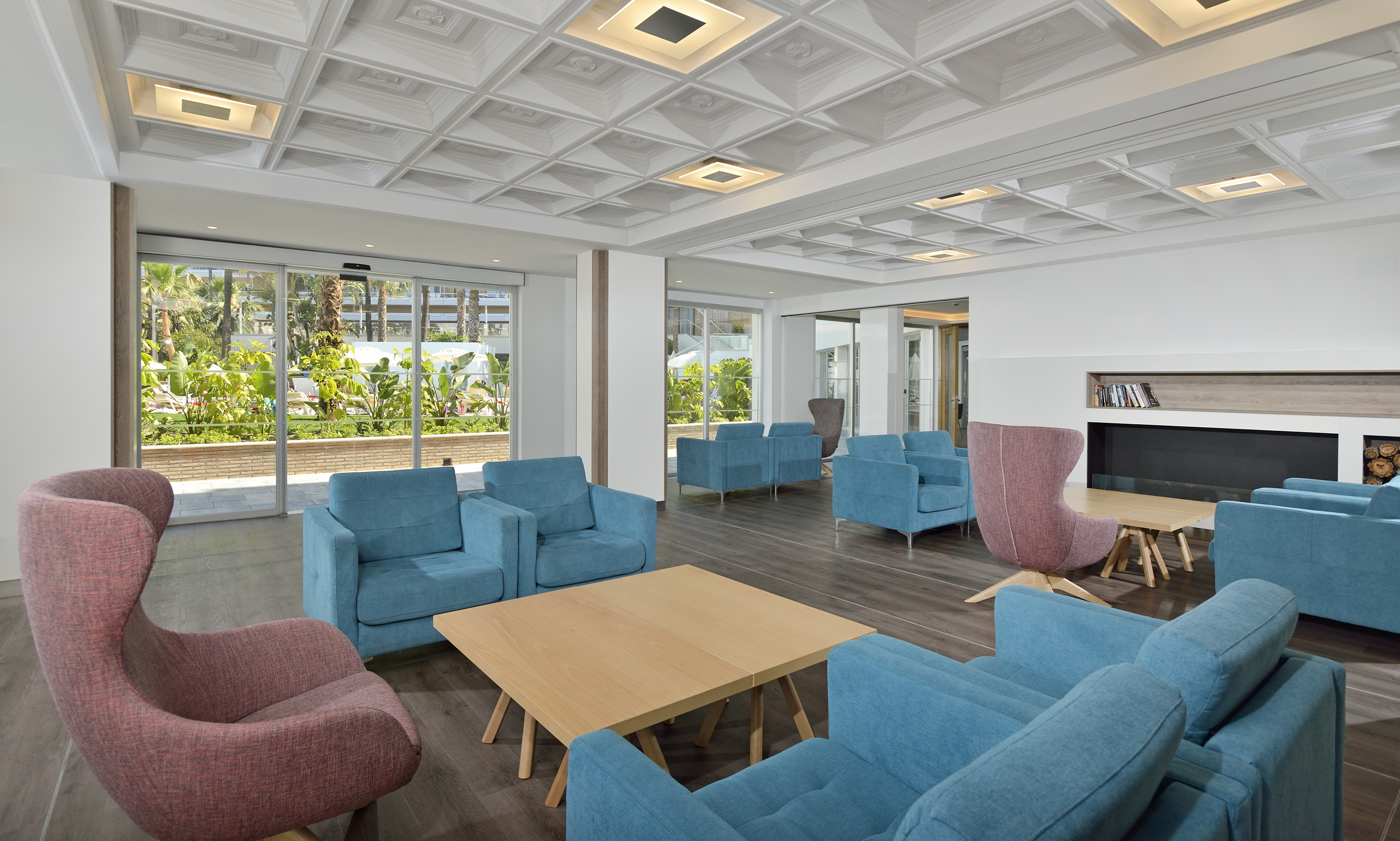 Modern hotel lounge with blue sofas, pink armchair, and tropical garden view.