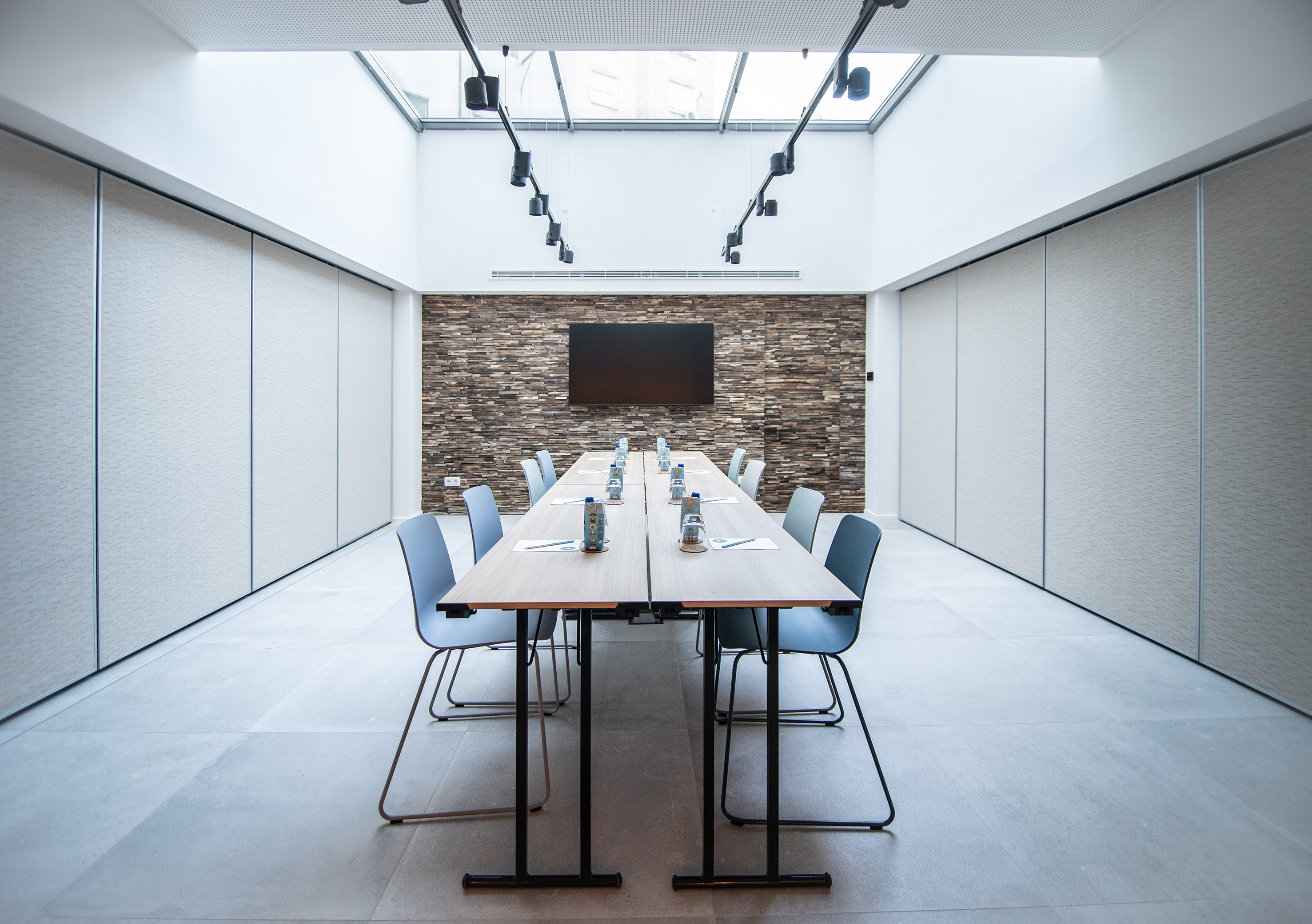 Modern meeting room with long table, blue chairs, screen, and stone wall.