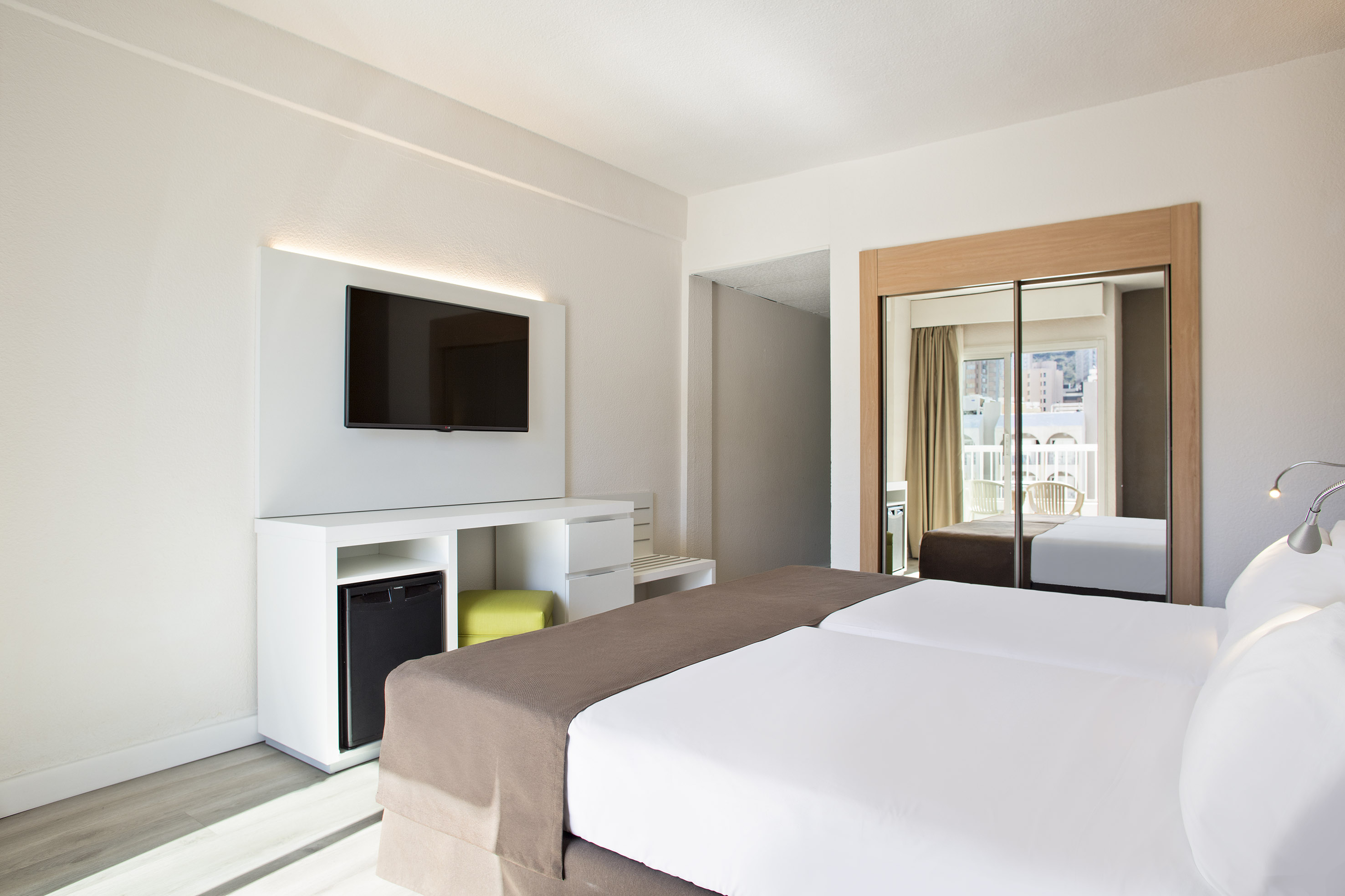 Bright modern hotel room with twin beds, TV, mini-fridge, and balcony view.
