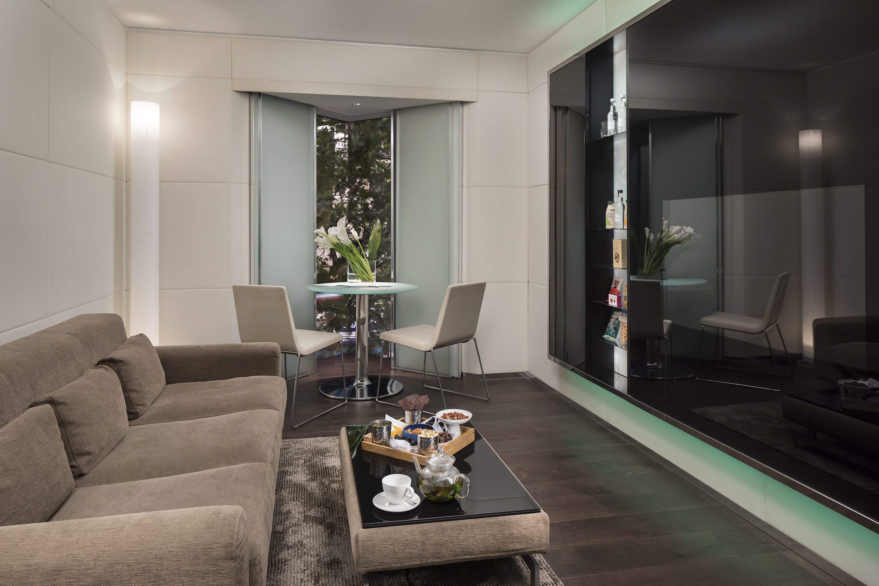 Modern hotel suite lounge with sofa, coffee table with tea, and window dining nook.