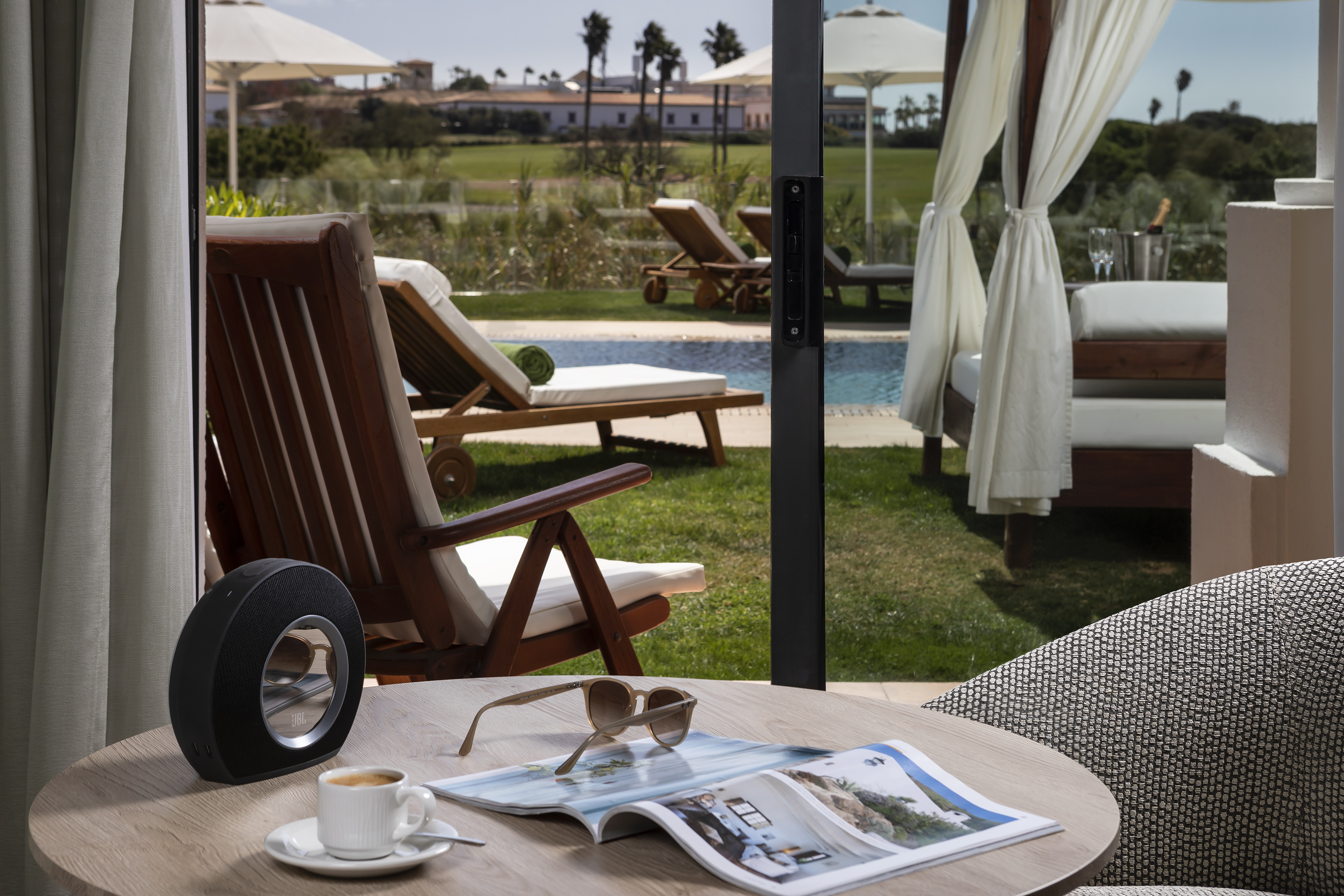 Resort patio with coffee, magazine, speaker, overlooking pool and lounge chairs.