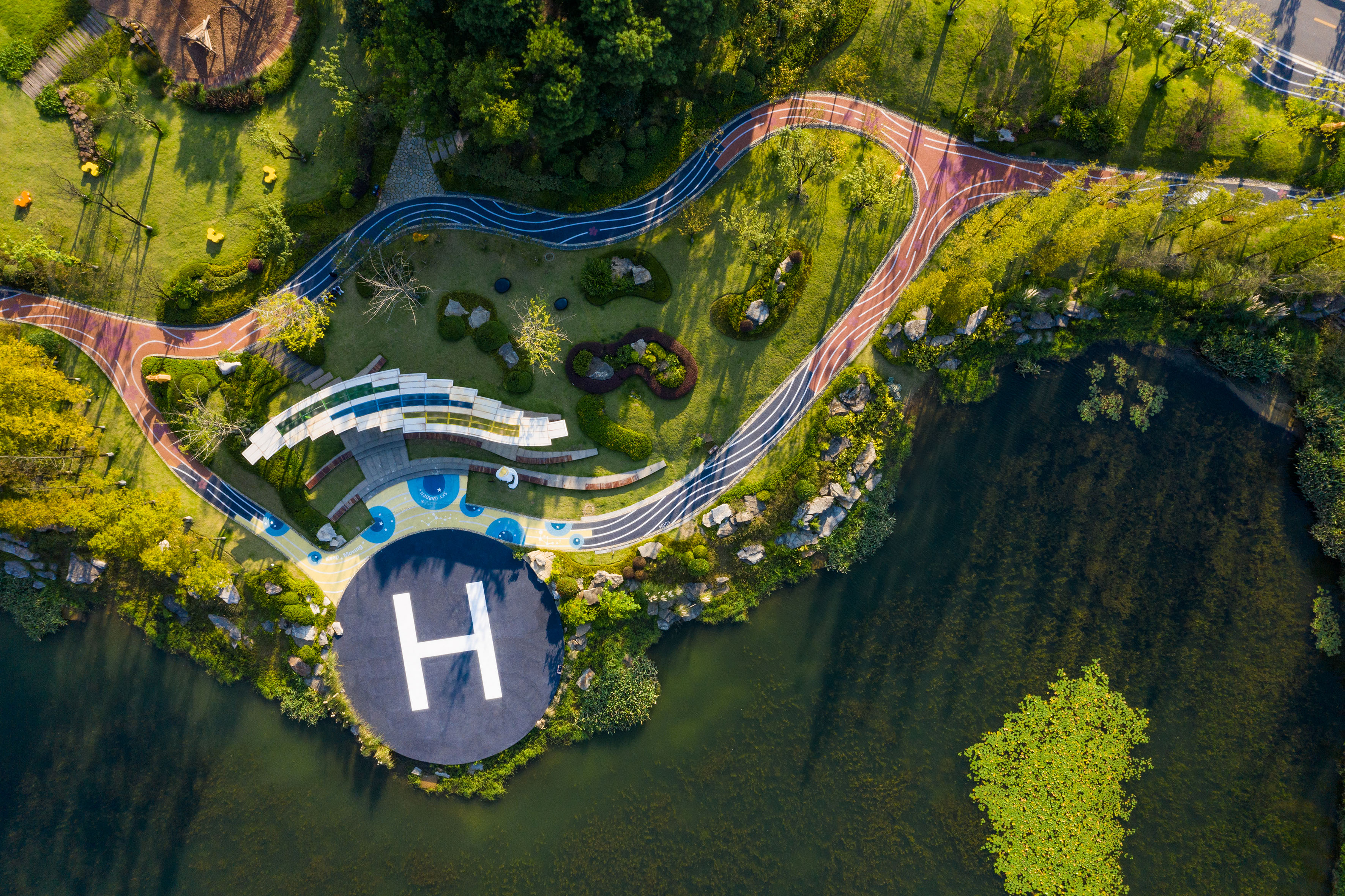 Aerial park: helipad by lake, winding tracks, green landscape, modern design.